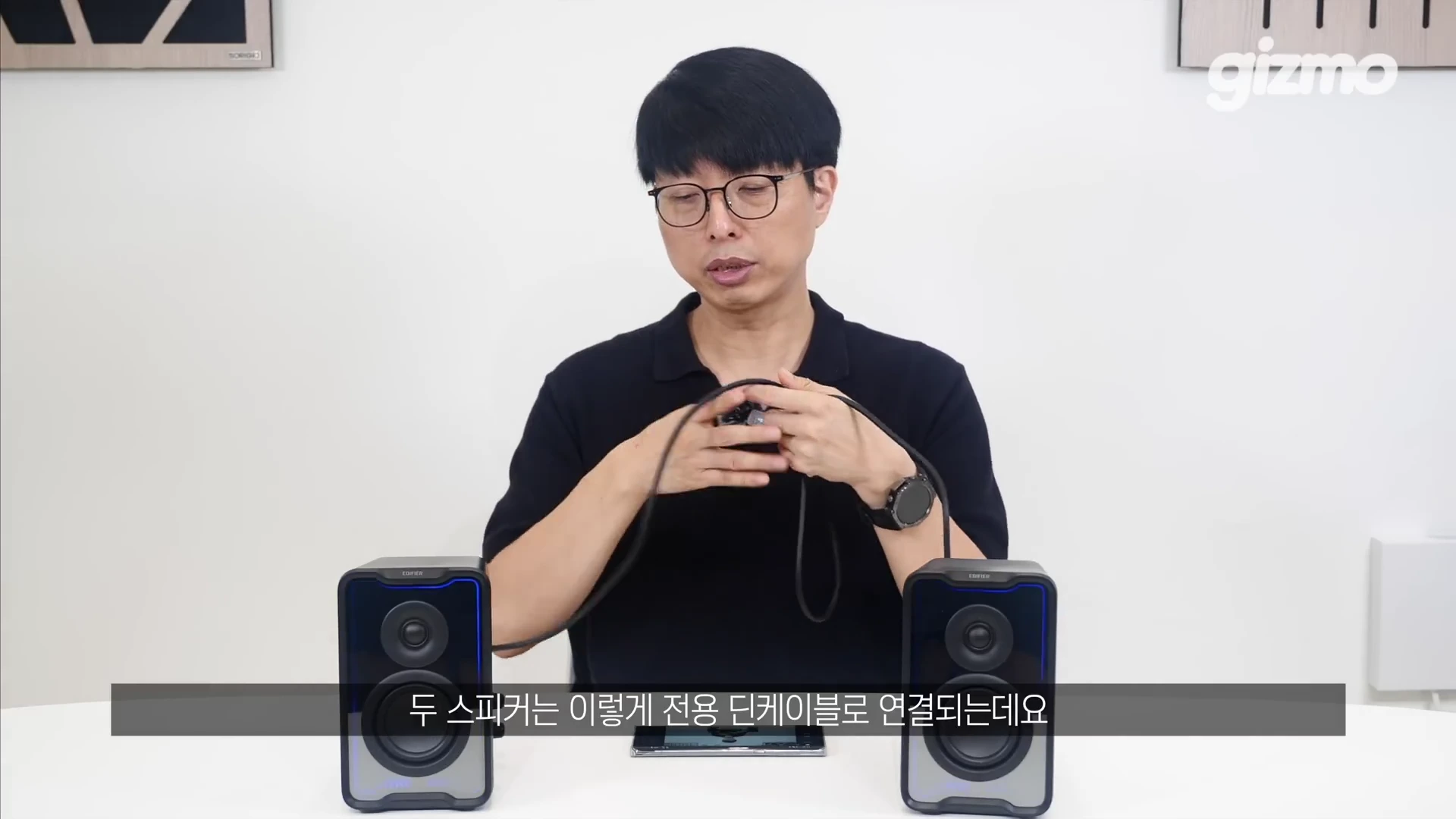 안경을 쓴 남성이 테이블 위에 놓인 두 개의 검은색 액티브 스피커 사이에서 전용 딘 케이블을 손에 들고 설명하는 모습
