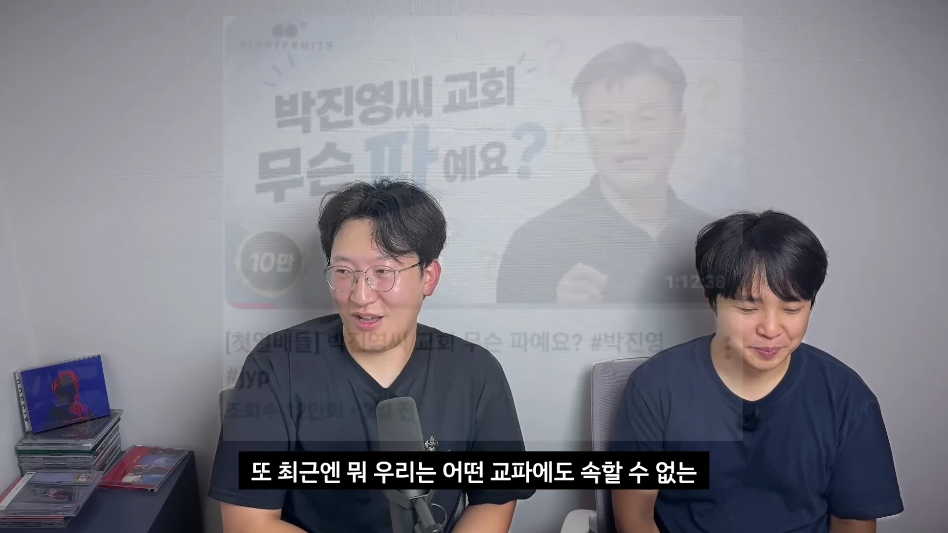 두 명의 남성이 책상 앞에 앉아 마이크를 앞에 두고 대화를 나누고 있으며, 뒤쪽 배경에는 '박진영 씨 교회 무슨 파예요?'라는 자막이 담긴 화면이 떠 있습니다.
