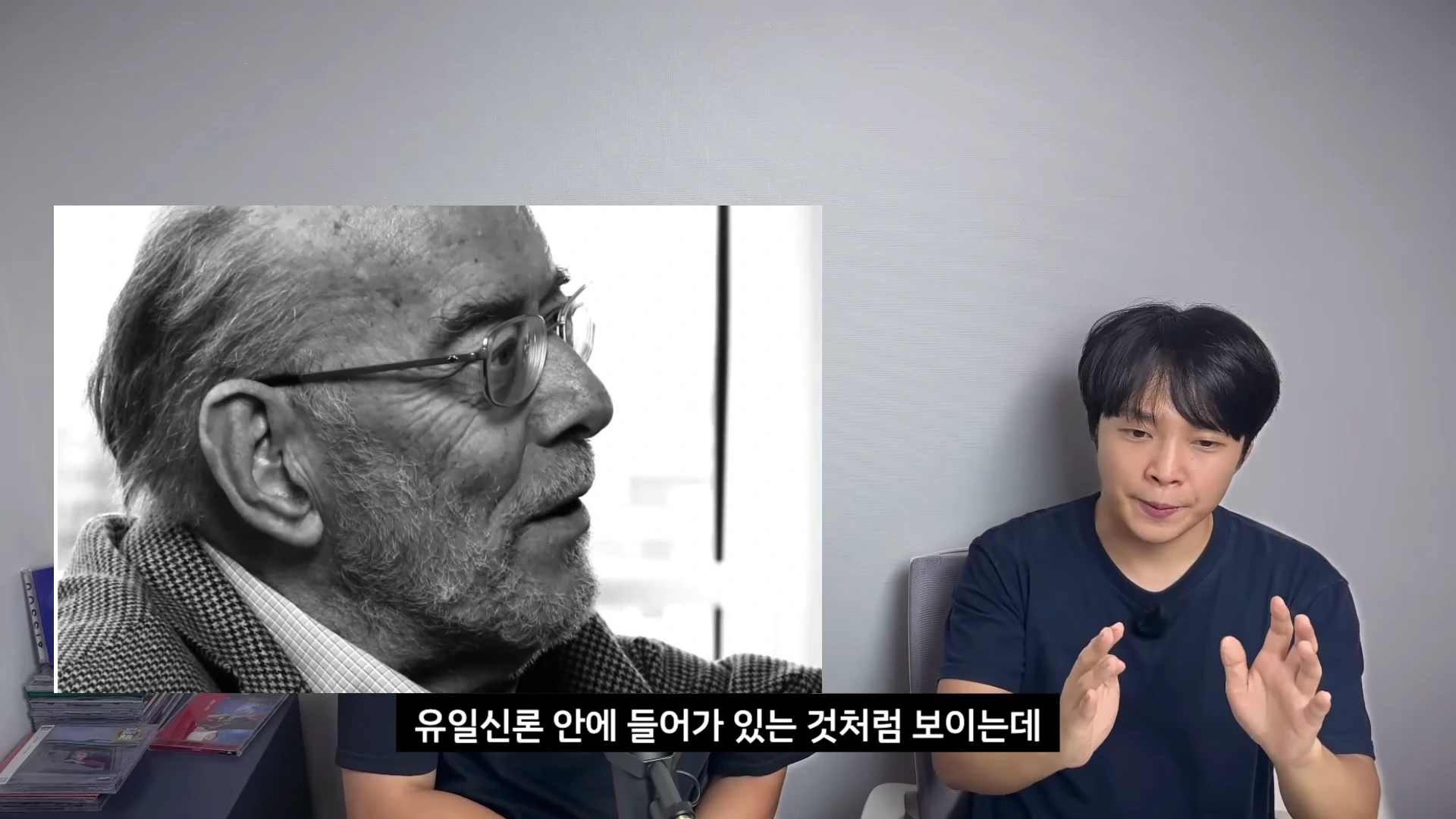 화면 오른쪽에는 검은색 티셔츠를 입은 남성이 앉아 이야기하고 있고, 왼쪽에는 흑백의 노신사 사진이 떠 있다.