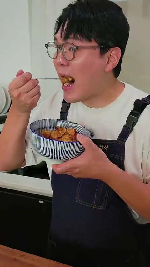 안경을 쓴 남성이 앞치마를 두르고 숟가락으로 덮밥을 한 입 떠먹는 모습을 담은 정면 영상 프레임입니다.