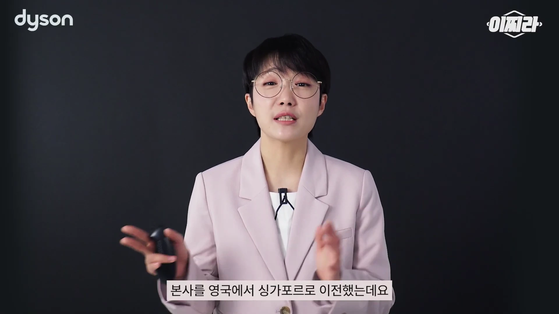 안경을 쓴 단발머리의 여성 발표자가 연한 분홍색 재킷을 입고 검은색 배경 앞에서 무언가를 설명하며 제스처를 취하고 있다.