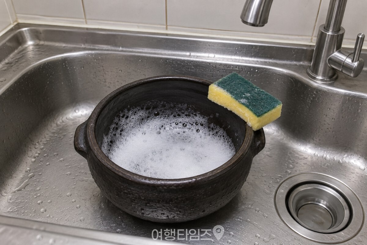 검은 회색 뚝배기 안쪽에 흰 세제 거품이 얇게 퍼져 있고, 노란 스펀지에 초록 연마면이 붙은 수세미가 뚝배기 테두리에 걸쳐져 있다. / 기사 이해를 돕기 위한 생성형 이미지