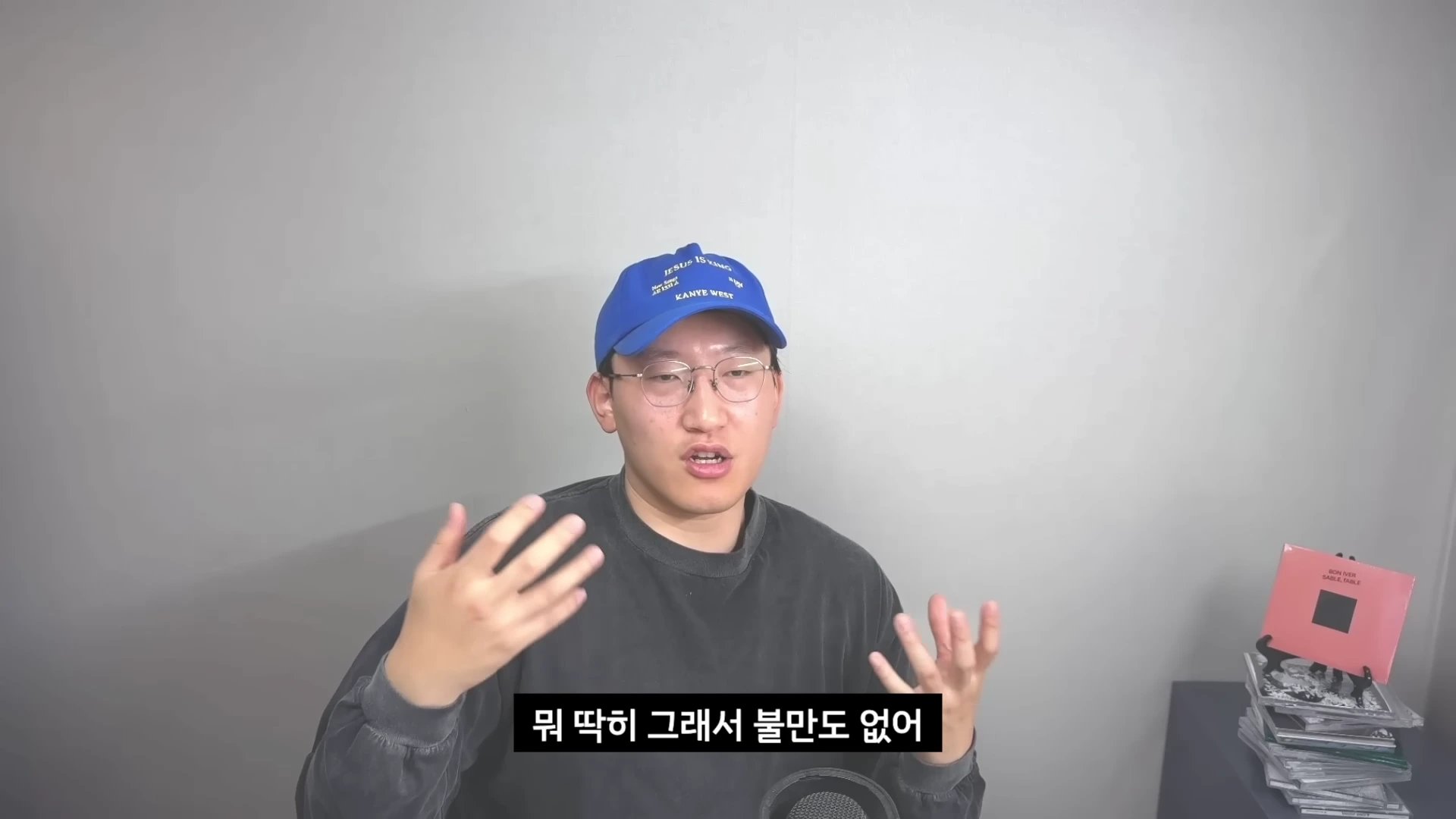 안경을 쓰고 파란색 볼캡을 쓴 남성이 회색 배경 앞에서 손짓하며 이야기를 하고 있으며 화면 하단에는 자막이 달려 있습니다.