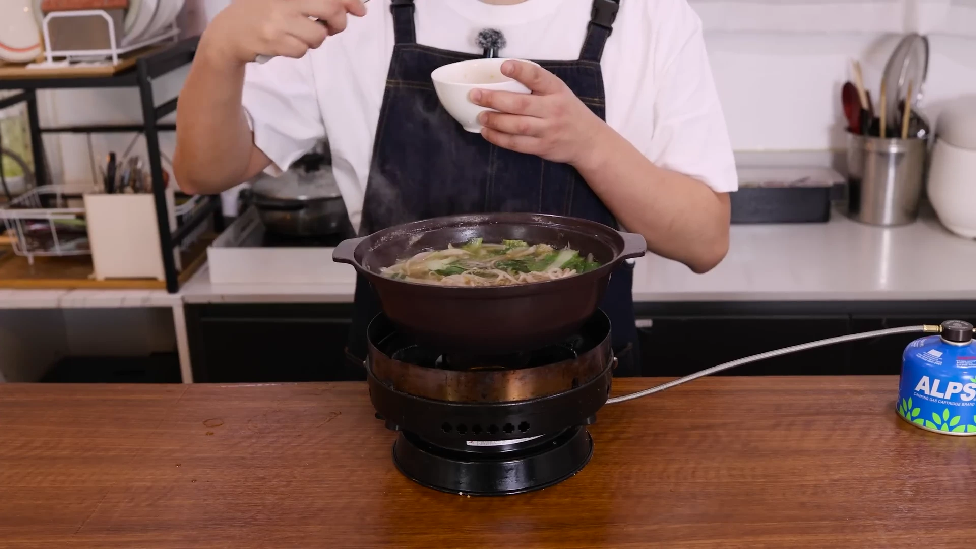 앞치마를 두른 사람이 휴대용 가스레인지 위의 뚝배기에 담긴 채소와 고기가 든 국물을 국자로 저으며 작은 그릇에 덜어내고 있다.