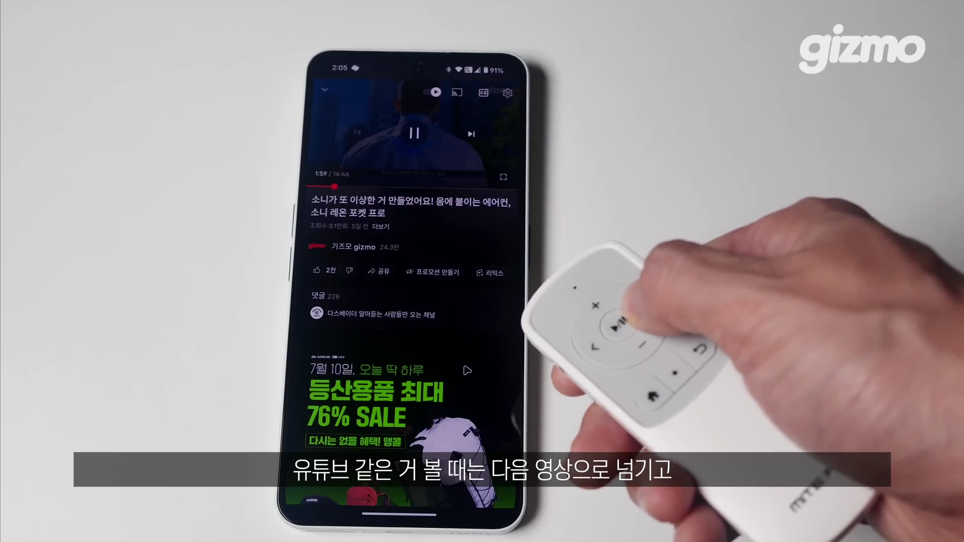 흰색 스마트폰 화면에 유튜브 영상이 재생되고 있으며, 손이 옆에서 미테르 리모컨을 사용하여 기기를 조작하고 있는 모습