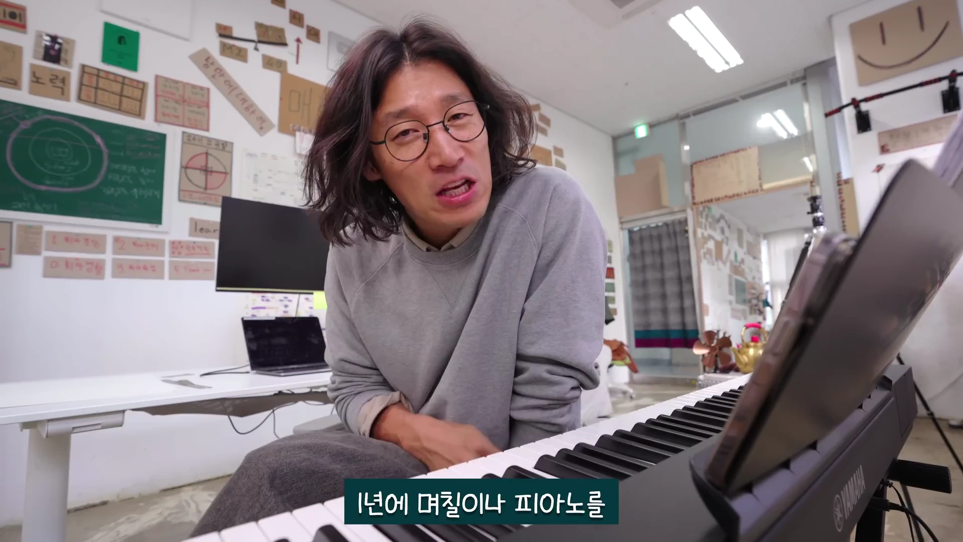 안경을 쓴 긴 머리의 남성이 피아노 앞에 앉아 카메라를 바라보며 이야기하고 있으며, 뒤쪽 벽면에는 다양한 메모와 글귀가 적힌 종이들이 붙어 있다.