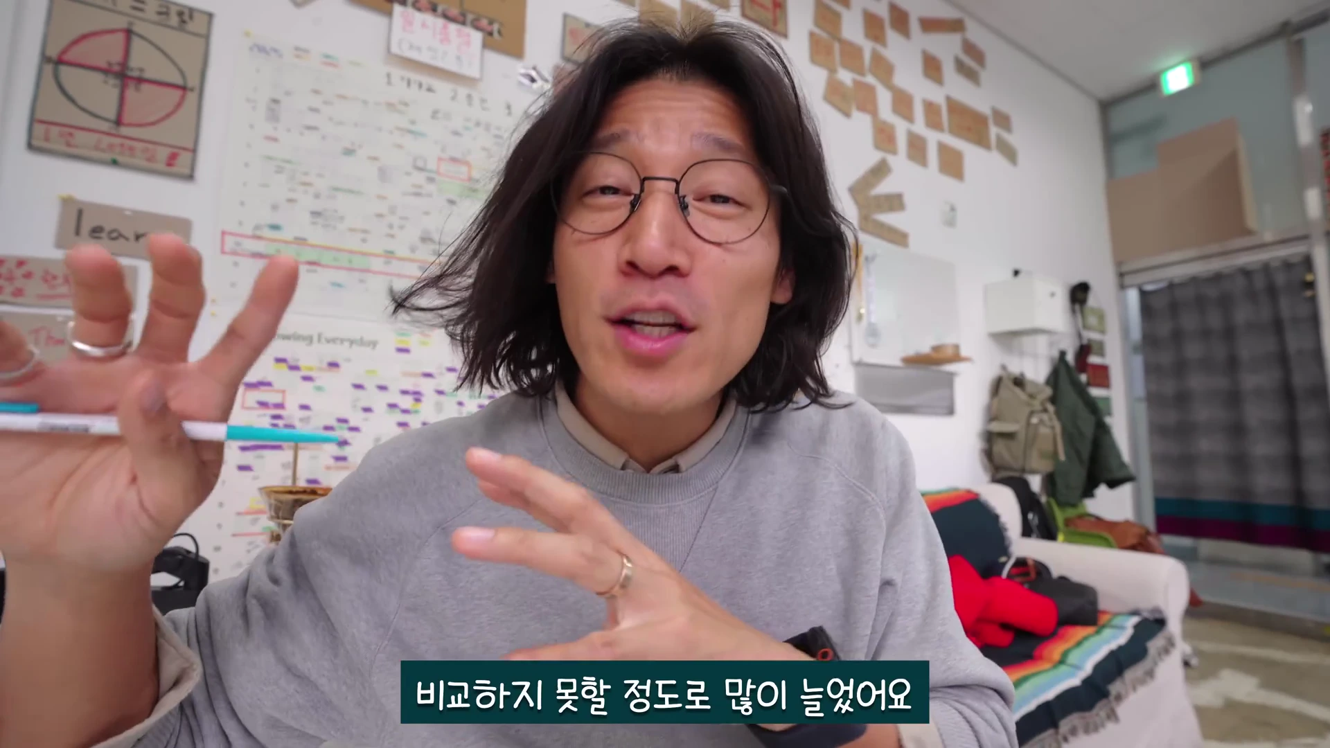 안경을 쓴 긴 머리 남성이 실내에서 카메라를 보며 손짓으로 설명하고 있고 뒤쪽 벽면에는 다양한 메모와 차트가 붙어 있습니다.