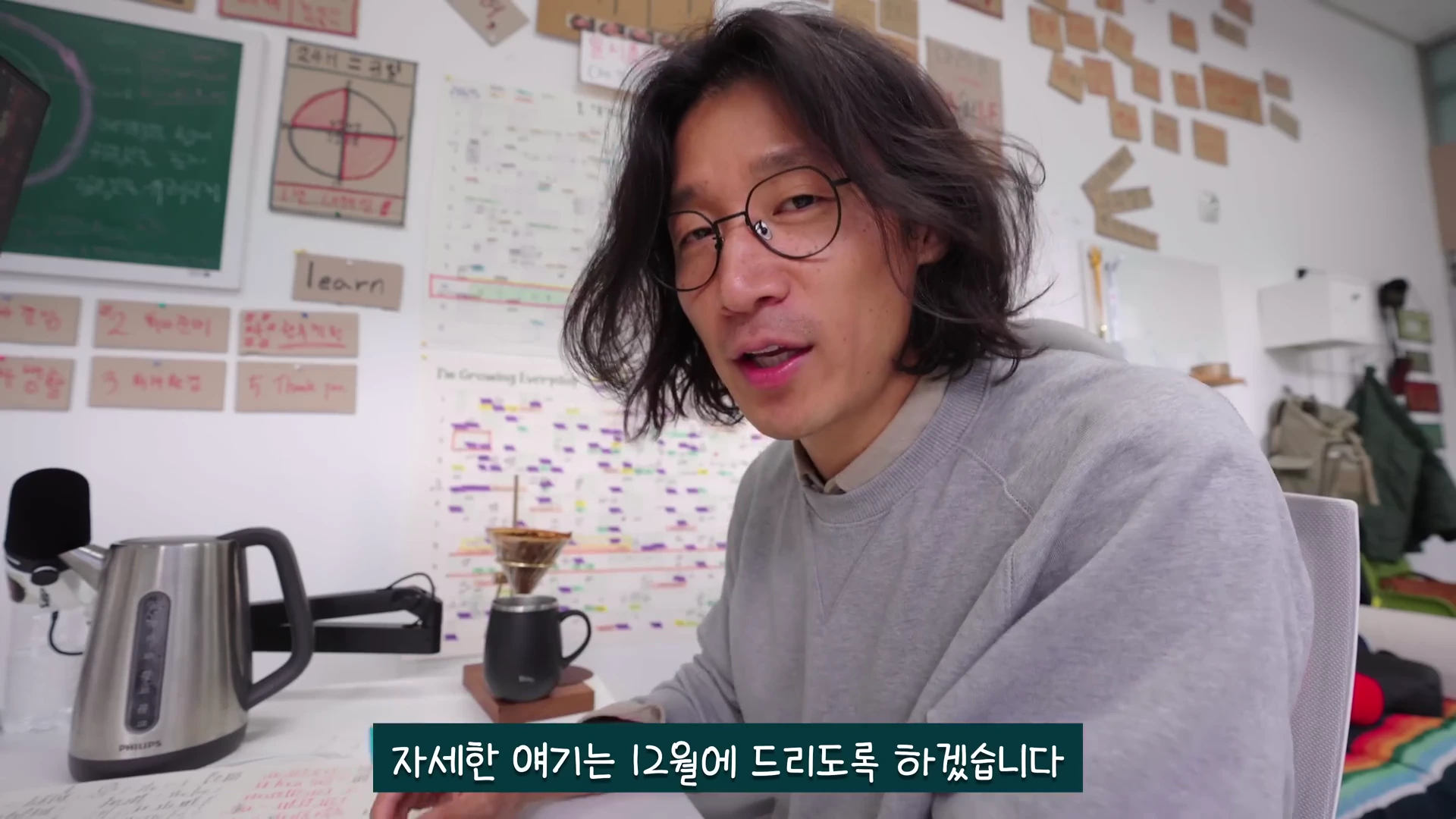 실내 작업실에서 안경을 쓴 중년 남성이 카메라를 보며 이야기하고 있고, 배경의 벽에는 다양한 메모와 차트가 붙어 있으며 책상 위에는 전기 주전자와 커피 도구가 놓여 있다.