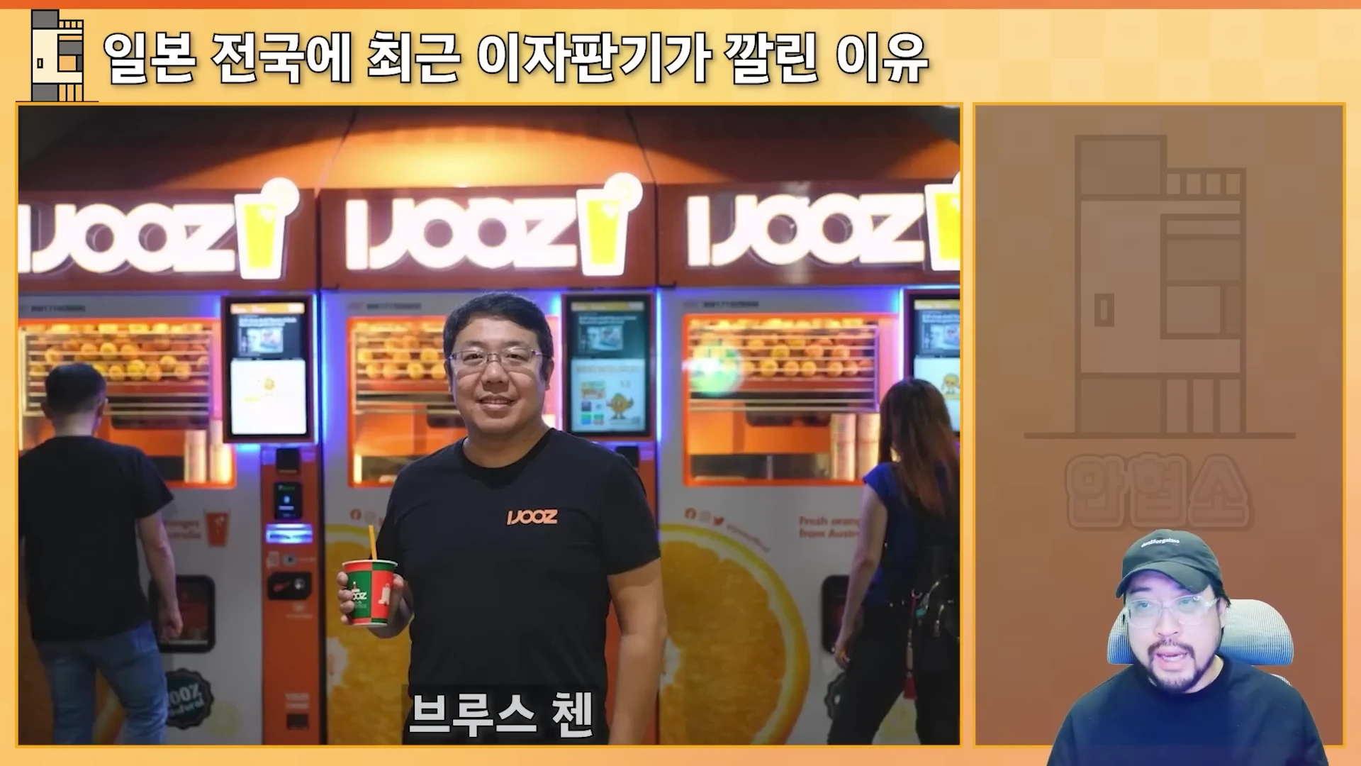 주황색 오렌지 착즙 자판기들이 나란히 놓인 실내를 배경으로, 검은색 티셔츠를 입은 남성이 주스 컵을 들고 서 있고, 우측 하단에는 모자를 쓴 진행자가 화면을 보고 말하는 모습이 담긴 영상 캡처 화면입니다.