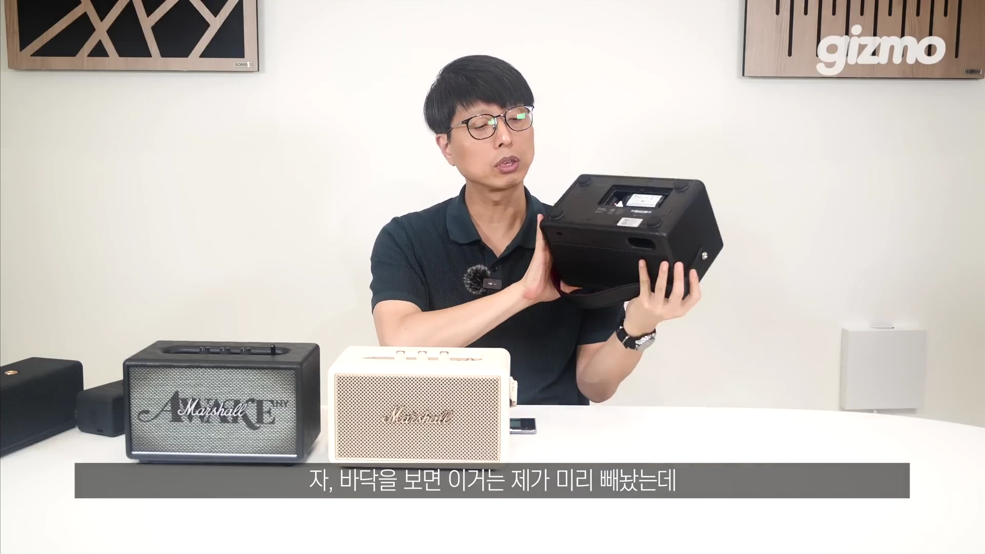 안경을 쓴 남성이 마샬 포터블 스피커의 바닥면을 들어 보이며 배터리 구조를 설명하고 있고, 앞쪽에는 다른 마샬 스피커 두 대가 놓여 있는 모습