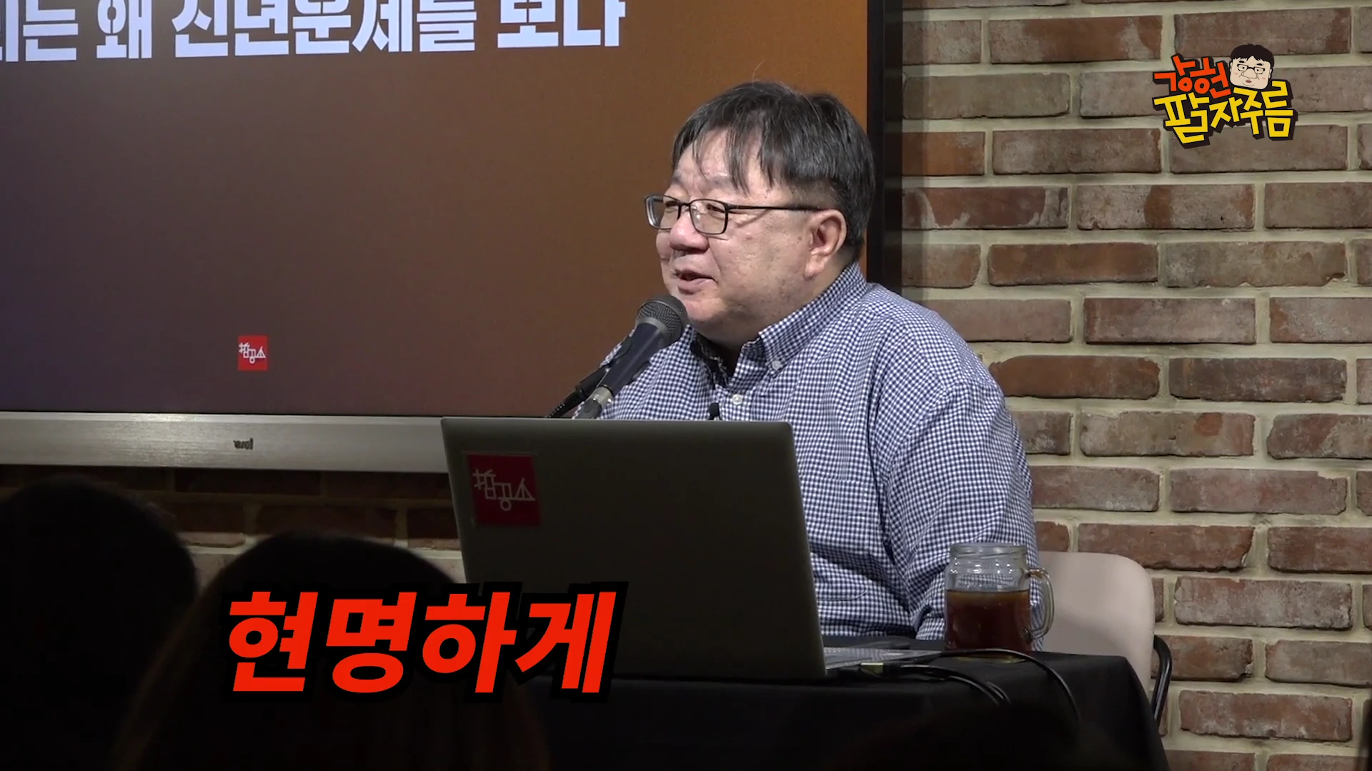 안경을 쓴 남성이 마이크 앞에 앉아 노트북을 보며 이야기하고 있는 스튜디오 장면