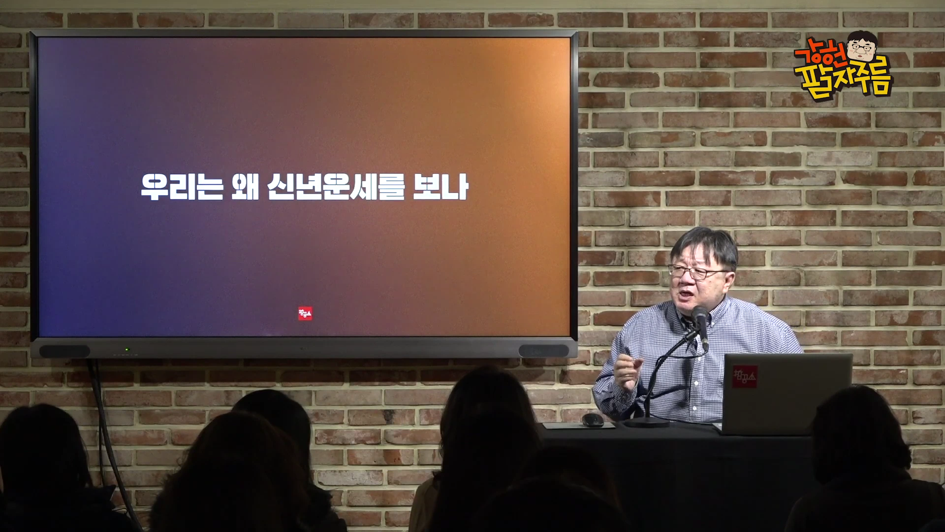 강연자로 보이는 한 남성이 벽돌 벽 배경의 스튜디오에서 '우리는 왜 신년운세를 보나'라는 제목이 띄워진 대형 화면을 앞에 두고 앉아 청중들을 향해 이야기하고 있다.