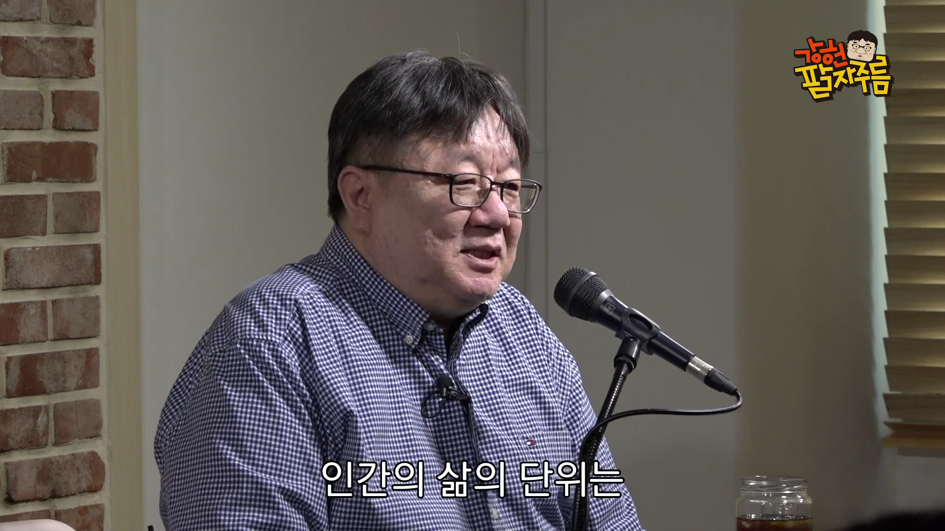 안경을 쓴 중년 남성이 벽돌 벽을 배경으로 마이크 앞에 앉아 이야기를 나누고 있는 모습이 담긴 스튜디오 영상 캡처 화면입니다.