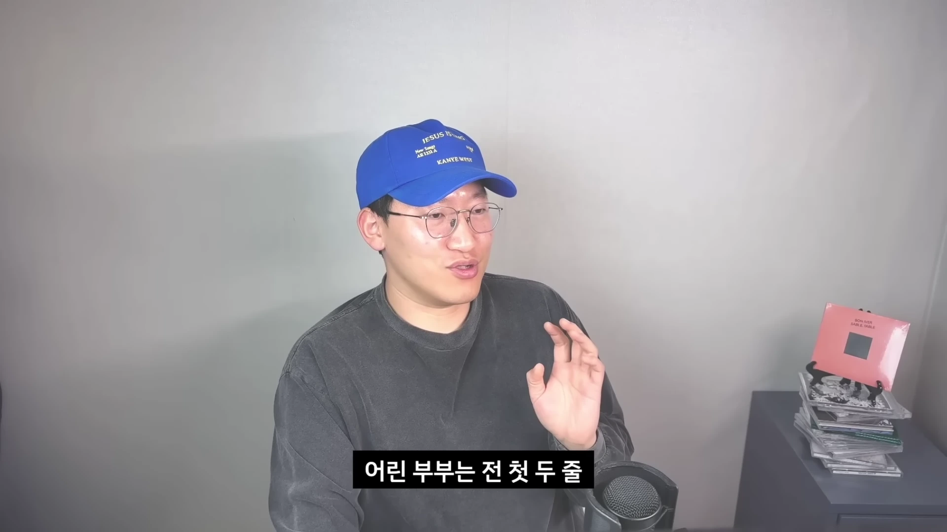 파란색 야구 모자와 안경을 착용한 남성이 회색 배경 앞에서 마이크를 앞에 두고 이야기를 나누고 있다.