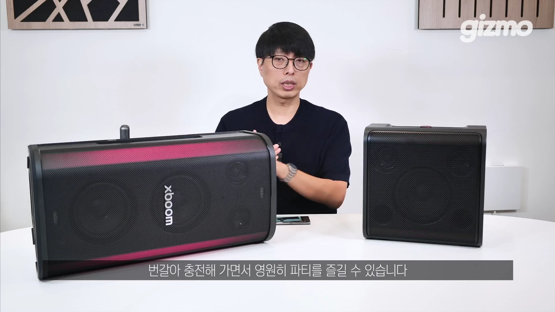 안경을 쓴 남성이 테이블 위에 놓인 두 대의 검은색 스피커 양옆에서 제스처를 하며 설명하는 모습