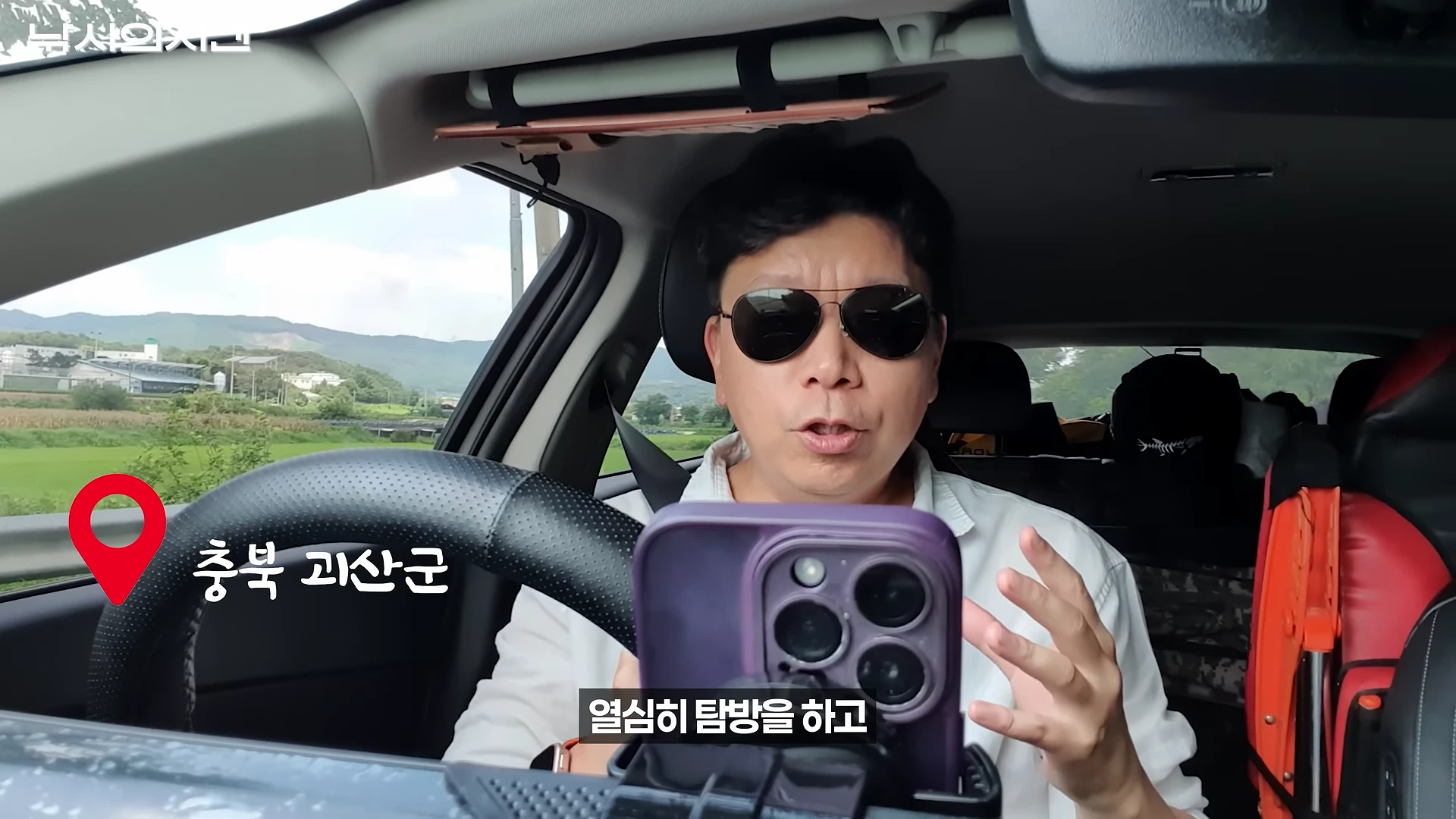 차량 운전석에 앉은 남성이 선글라스를 쓰고 스마트폰을 바라보며 이야기하고 있으며, 화면에는 '충북 괴산군'이라는 문구와 위치 표시 아이콘이 떠 있습니다.