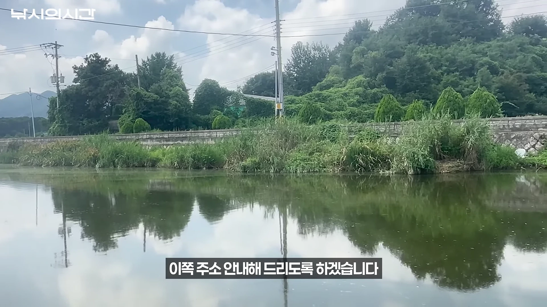 잔잔한 물가 주변으로 우거진 수풀과 전신주가 보이고 하단에 자막이 있는 낚시터 풍경