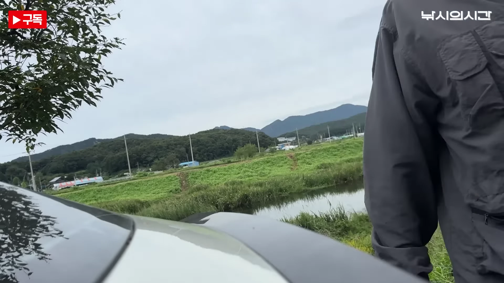 금당천 주변의 탁 트인 야외 낚시터 풍경을 배경으로 촬영자가 서 있는 모습이 담긴 영상 캡처 화면입니다. 화면 오른쪽에는 낚시 조끼를 입은 인물의 뒷모습이 보이고, 앞쪽으로는 풀이 우거진 하천과 산이 펼쳐져 있습니다.