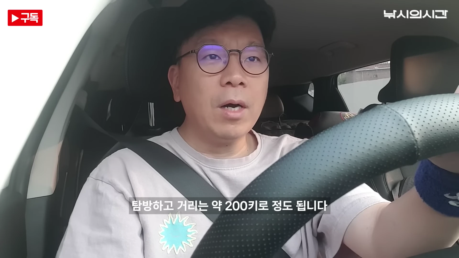 안경을 쓴 중년 남성이 운전석에서 정면을 바라보며 이야기하고 있는 자동차 내부 모습