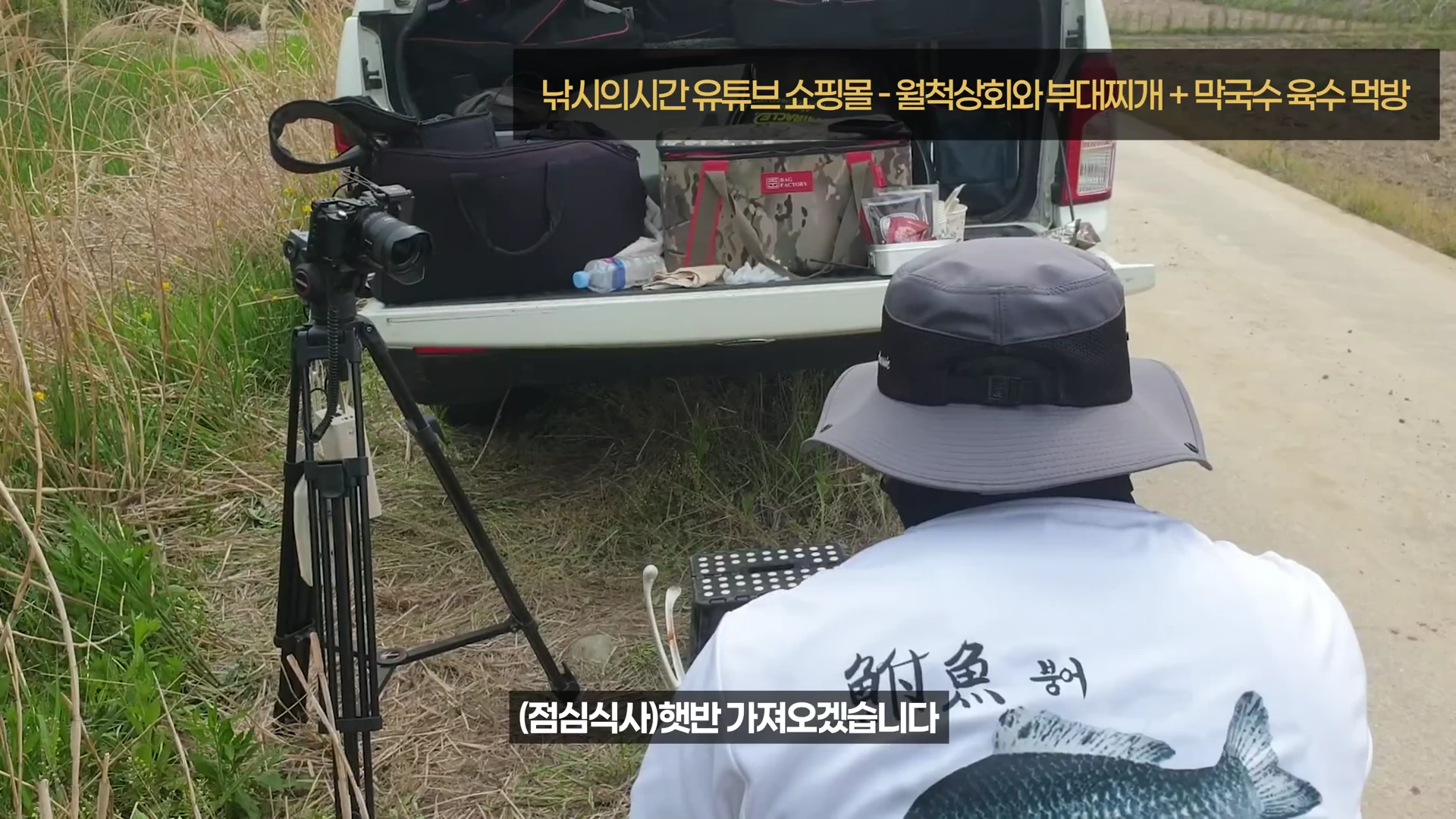 낚시터 노지에서 등 뒤에 붕어 그림이 그려진 흰색 티셔츠와 모자를 착용한 남성이 촬영 장비를 세워둔 채 야외 식사를 준비하고 있는 모습