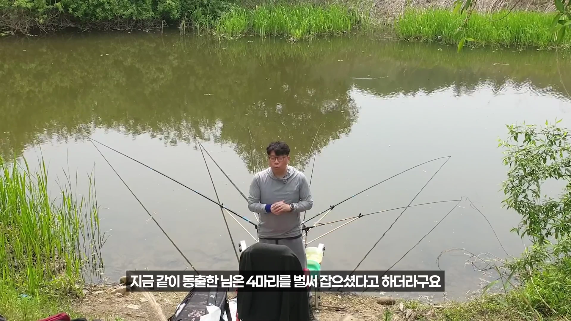 강가에서 회색 긴팔 티셔츠를 입은 남성이 낚싯대를 여러 대 펼쳐놓고 서 있는 모습
