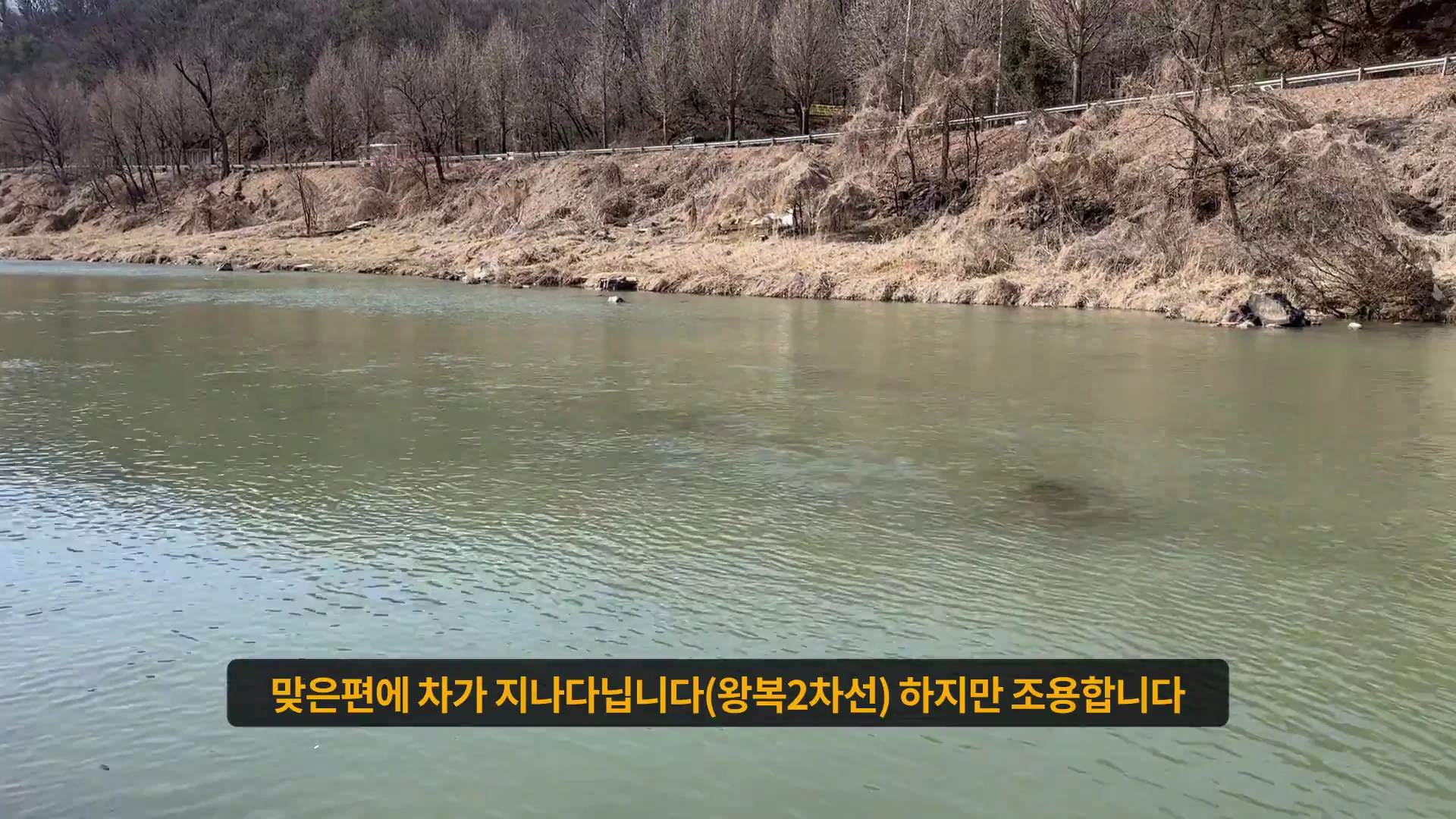 흐린 물색의 강과 그 뒤로 마른 풀이 덮인 언덕, 그리고 위쪽으로 도로가 보이는 풍경 사진이며 화면 하단에는 노란색 자막이 배치되어 있습니다.