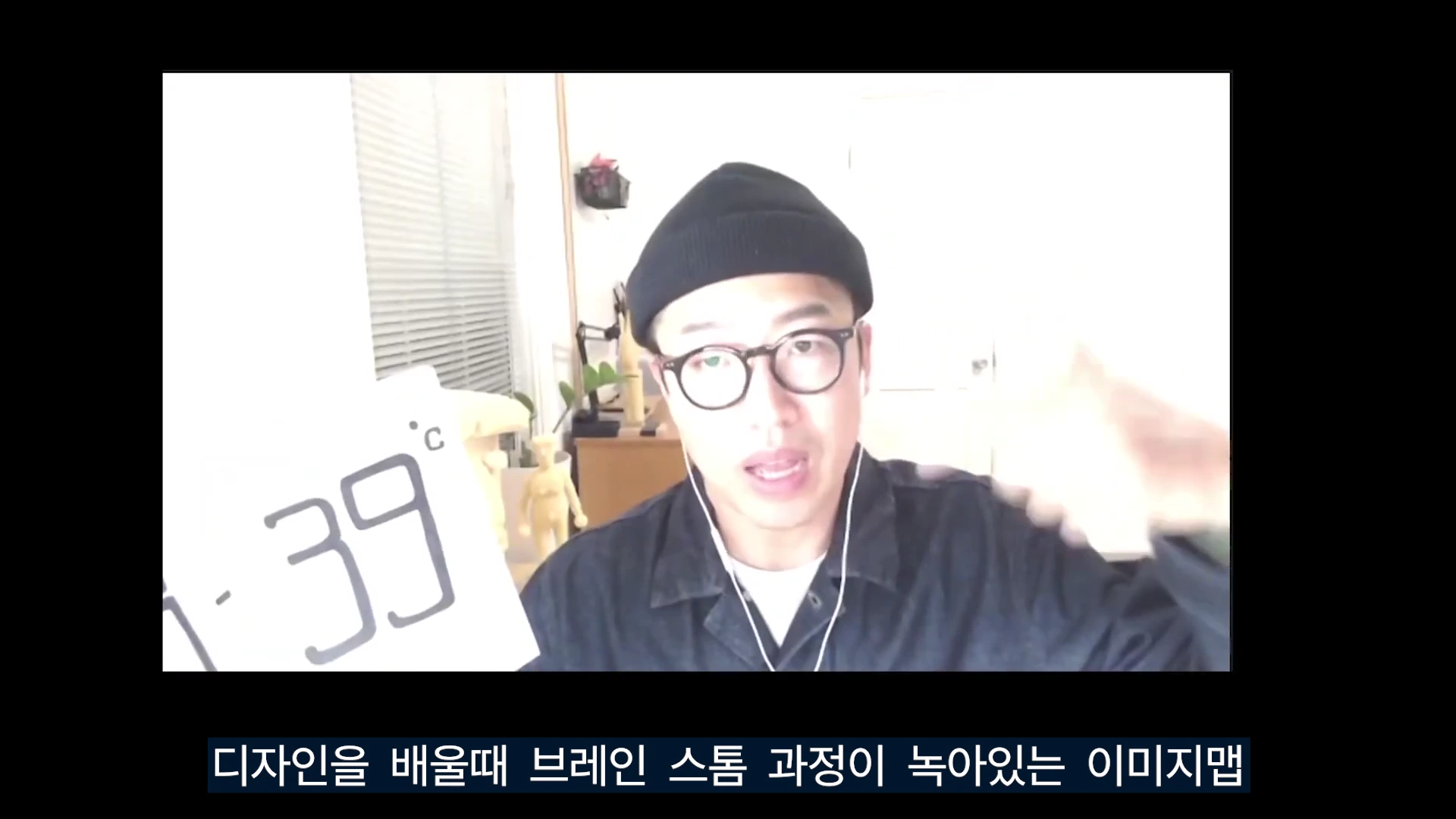 비니를 쓰고 안경을 쓴 남성이 웹캠 영상 속에서 손짓하며 이야기하고 있고, 화면 왼쪽에는 '38 39°C'라는 글자가 적힌 종이가 배치되어 있다.