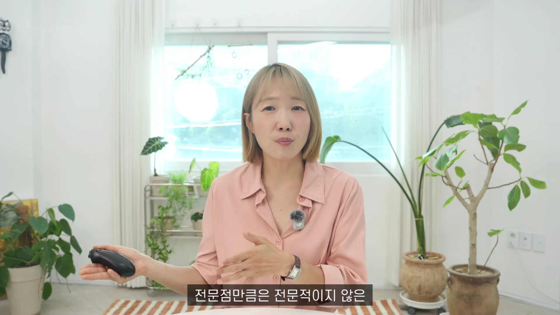 A woman with short, blonde hair wearing a light pink shirt is speaking to the camera in a room filled with potted plants, holding a small remote control in her right hand.