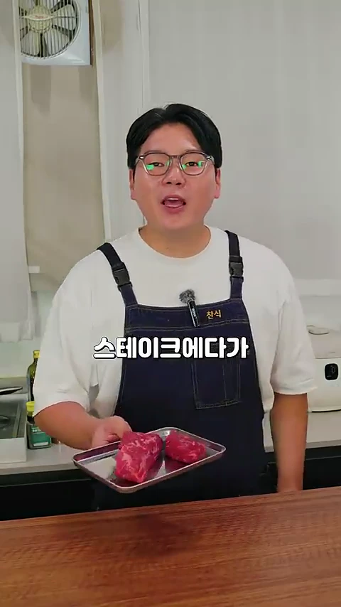 안경을 쓴 남성이 앞치마를 두르고 스테이크용 생고기가 담긴 쟁반을 들고 주방에서 이야기하고 있다.