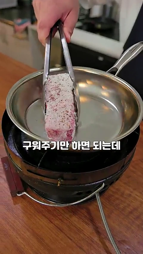 집게로 프리마 가루가 묻은 스테이크 고기를 들어 프라이팬 위에 올리는 모습이 담긴 조리 영상의 한 장면입니다.