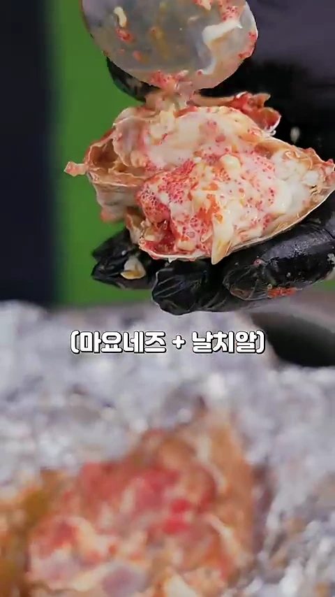 검은색 위생 장갑을 낀 손이 마요네즈와 날치알이 듬뿍 섞인 소스를 구운 꽃게 등딱지 위에 숟가락으로 올리는 모습.