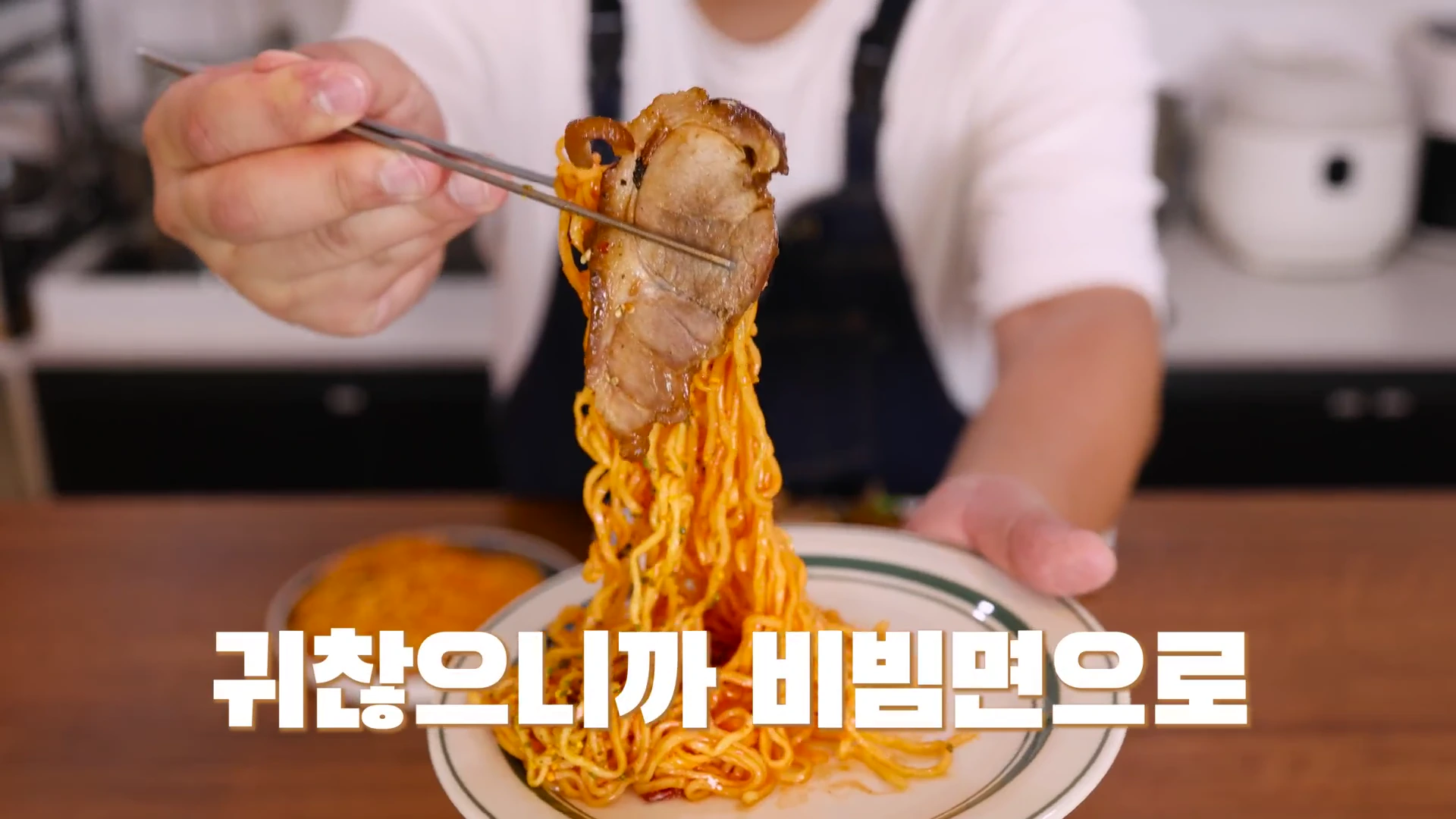 흰색 티셔츠와 앞치마를 입은 사람이 젓가락으로 비빔면 한 점과 그 위에 올린 족발 고기를 들어 카메라 쪽으로 내밀고 있는 모습.