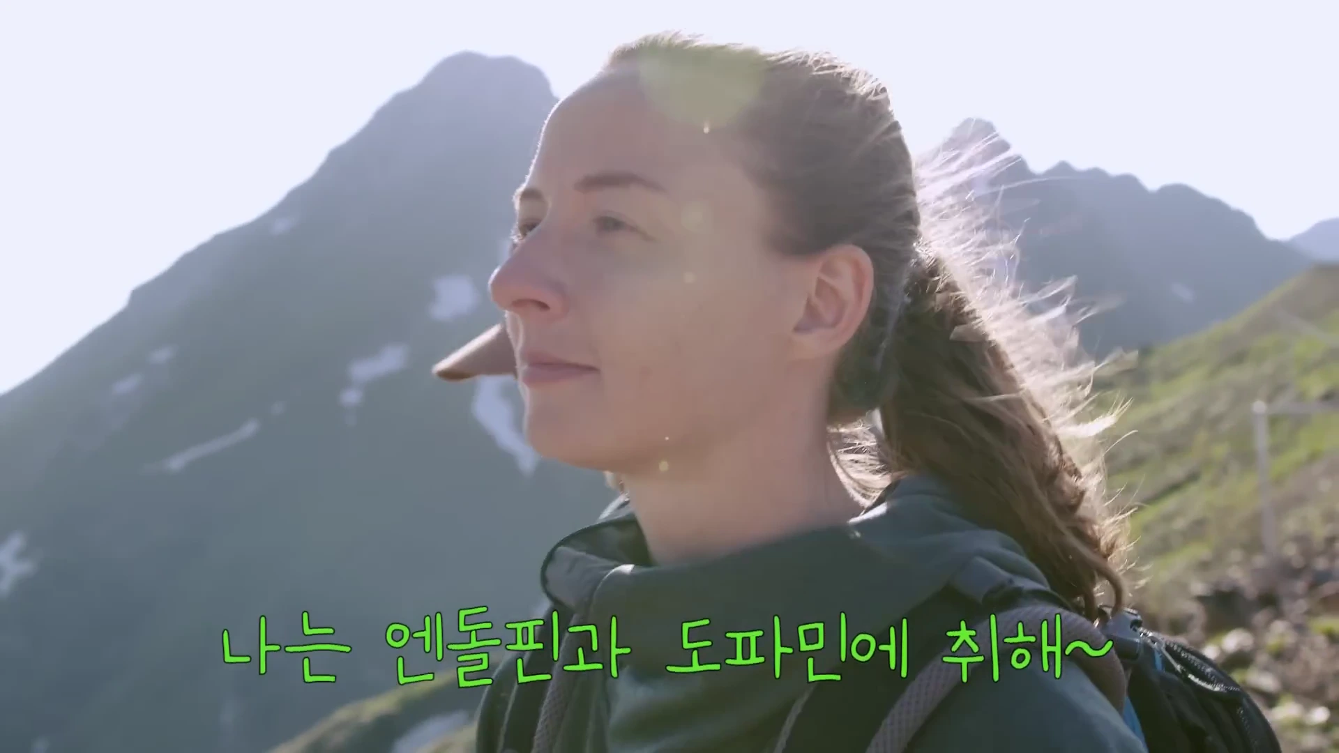 산맥을 배경으로 백팩을 멘 여성이 맑은 하늘 아래에서 정면을 응시하며 미소 짓고 있는 영상의 한 장면입니다.
