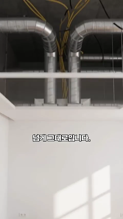 공사 중인 아파트 천장 내부의 모습으로, 콘크리트 슬래브 아래에 설치된 은색 금속 덕트와 노란색 전선들이 복잡하게 얽혀 있고 그 아래로 텍스트가 적힌 화면.