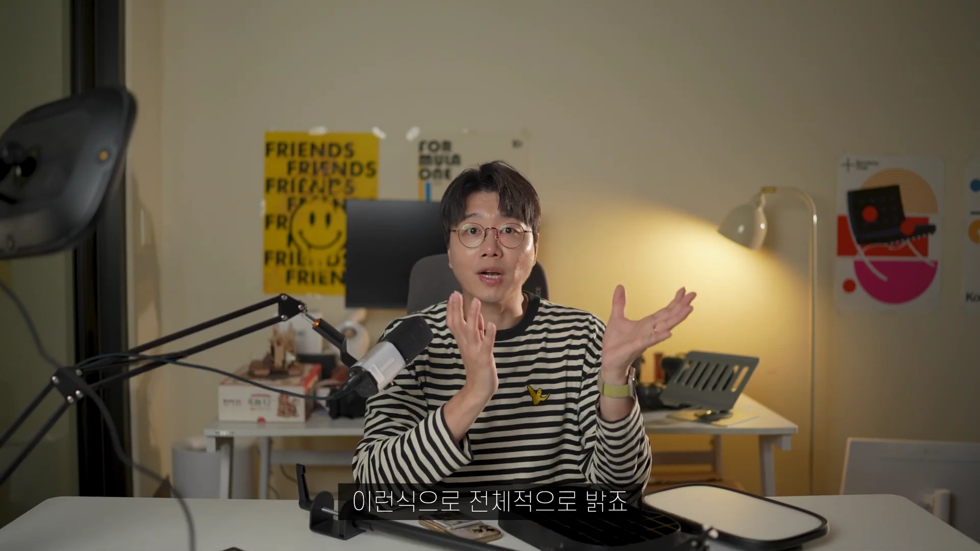 안경을 쓴 남성이 책상 앞에 앉아 양손을 들어 설명하며 말하고 있는 모습이 담긴 영상 캡처 화면이며, 왼편에는 조명 장비와 마이크가 배치되어 있다.