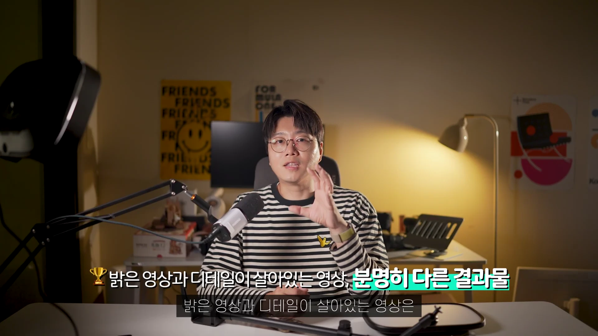 안경을 쓴 남성이 책상 앞에 앉아 제스처를 하며 이야기하고 있으며, 그 주변으로 조명과 마이크 등 촬영 장비가 배치된 모습이 담긴 영상 캡처 화면입니다.