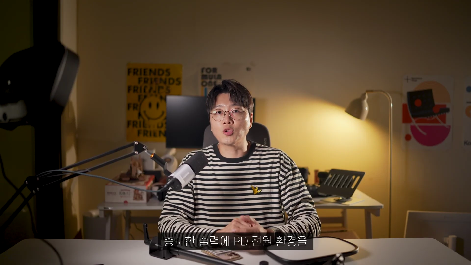 안경을 쓴 남성이 스트라이프 티셔츠를 입고 책상 앞에 앉아 이야기하는 모습의 스튜디오 촬영 화면.