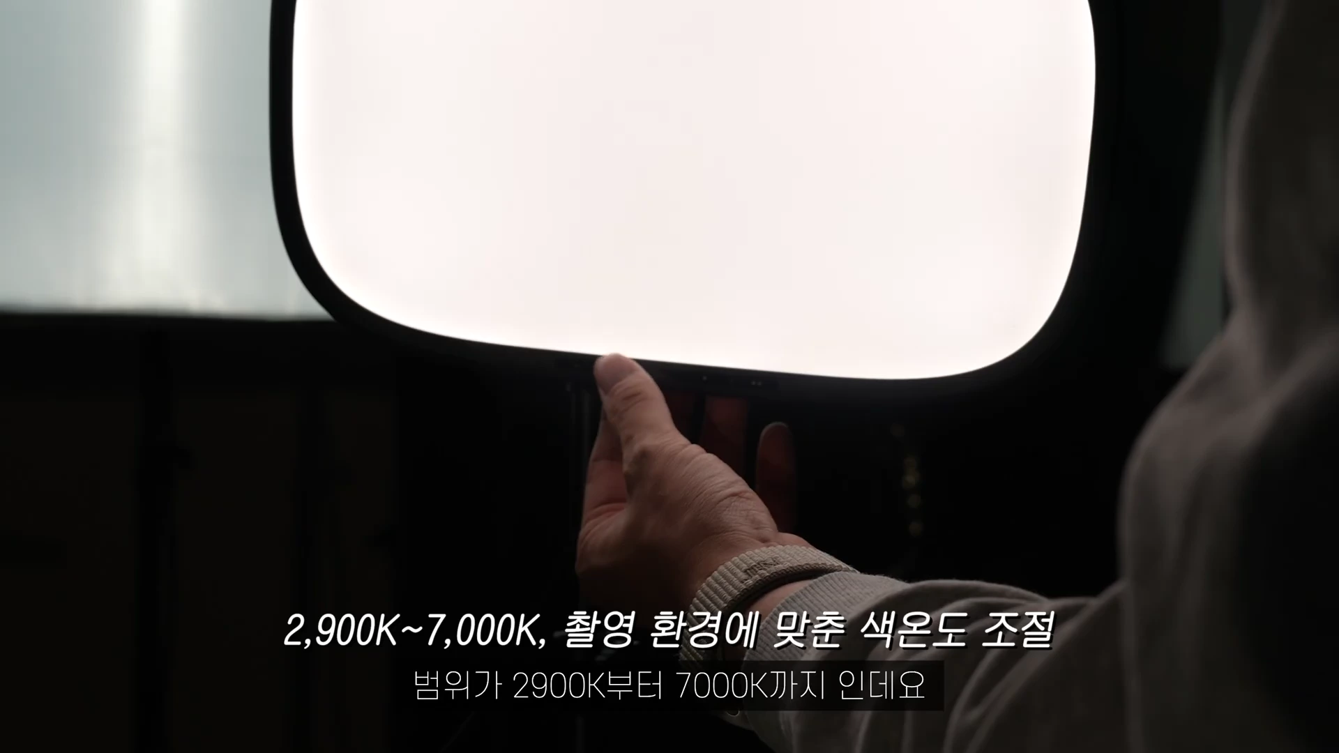 촬영 조명 하단부를 손으로 조절하는 모습이 담긴 클로즈업 샷으로, 화면 하단에는 색온도 조절 범위를 설명하는 자막이 함께 표시되어 있습니다.