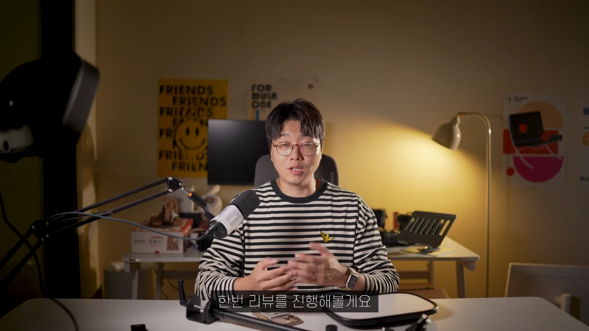 안경을 쓴 남성이 줄무늬 티셔츠를 입고 책상 앞에 앉아 카메라를 향해 말하고 있으며, 좌측에는 조명 장비가, 책상 위에는 부드러운 빛을 내는 조명 판과 마이크가 놓여 있다.