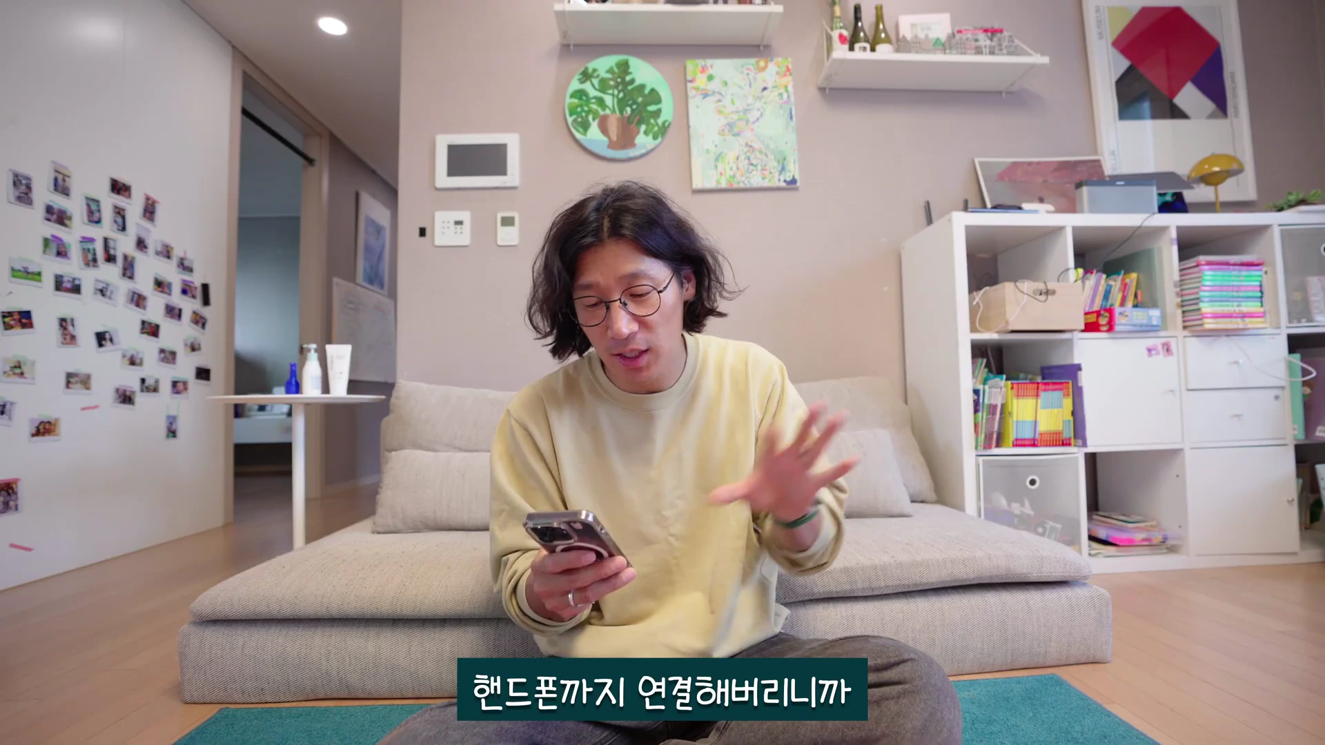안경을 쓴 남성이 거실 소파에 앉아 스마트폰을 보며 손짓으로 설명하고 있는 모습.