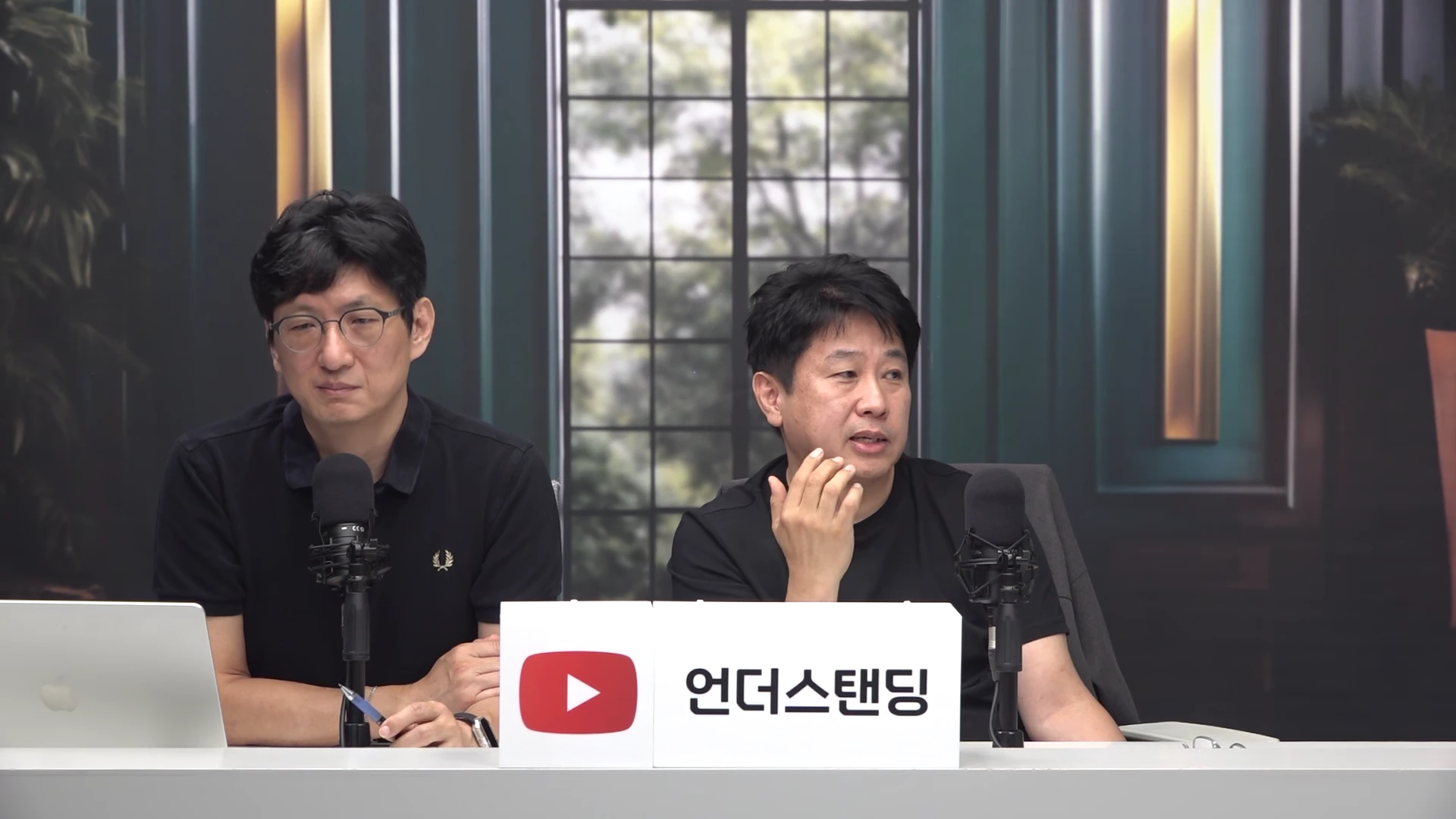 두 명의 남성이 검은색 옷을 입고 마이크가 설치된 책상 앞에 앉아 대화하고 있다.