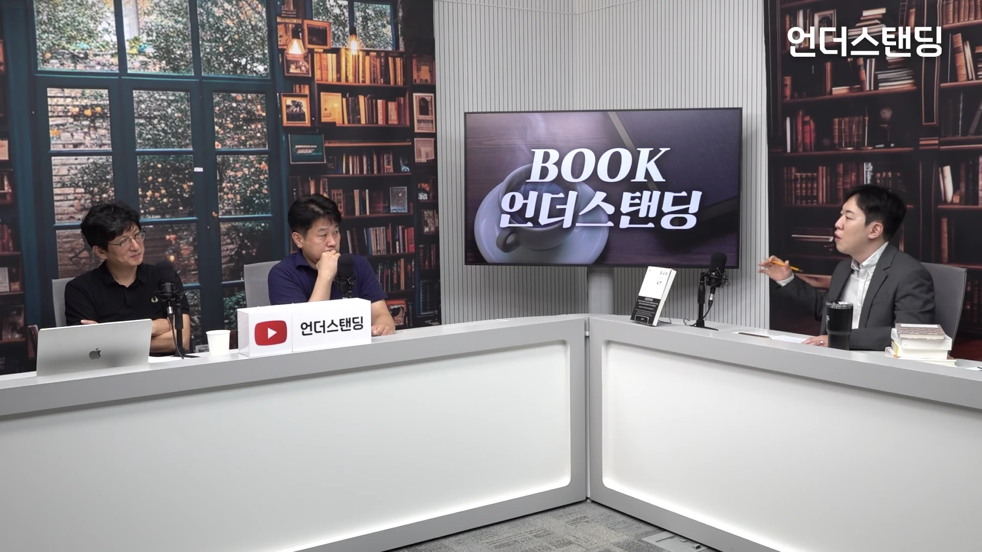 두 명의 출연자가 스튜디오에서 대담을 나누는 모습. 책상 위에 마이크와 책이 놓여 있고, 뒤쪽 벽에는 'BOOK 언더스탠딩'이라는 문구와 커피 잔 이미지가 담긴 대형 화면이 배치되어 있다.
