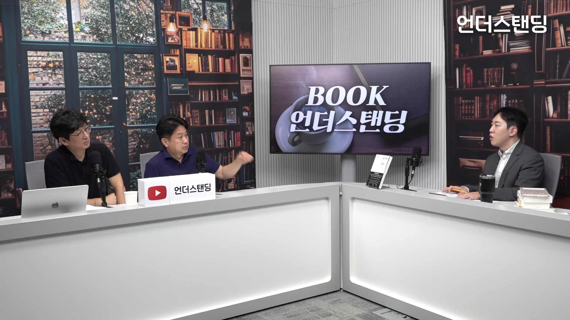 서재 배경의 스튜디오에서 세 명의 남성이 책상에 앉아 대화를 나누고 있는 모습으로, 중앙에 있는 남성이 손동작을 하며 설명하고 있고 화면에는 북 프로그램 로고가 송출되고 있습니다.