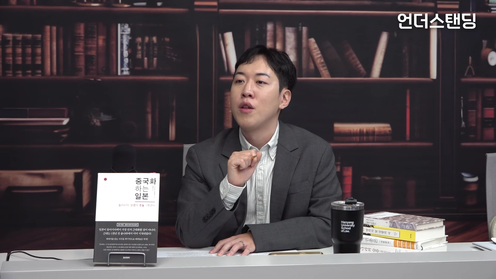 서재 배경 앞에서 정장을 입은 한 남성이 책상에 앉아 이야기하는 모습이며, 왼쪽에는 책이 세워져 있고 오른쪽에는 텀블러와 책이 쌓여 있다.