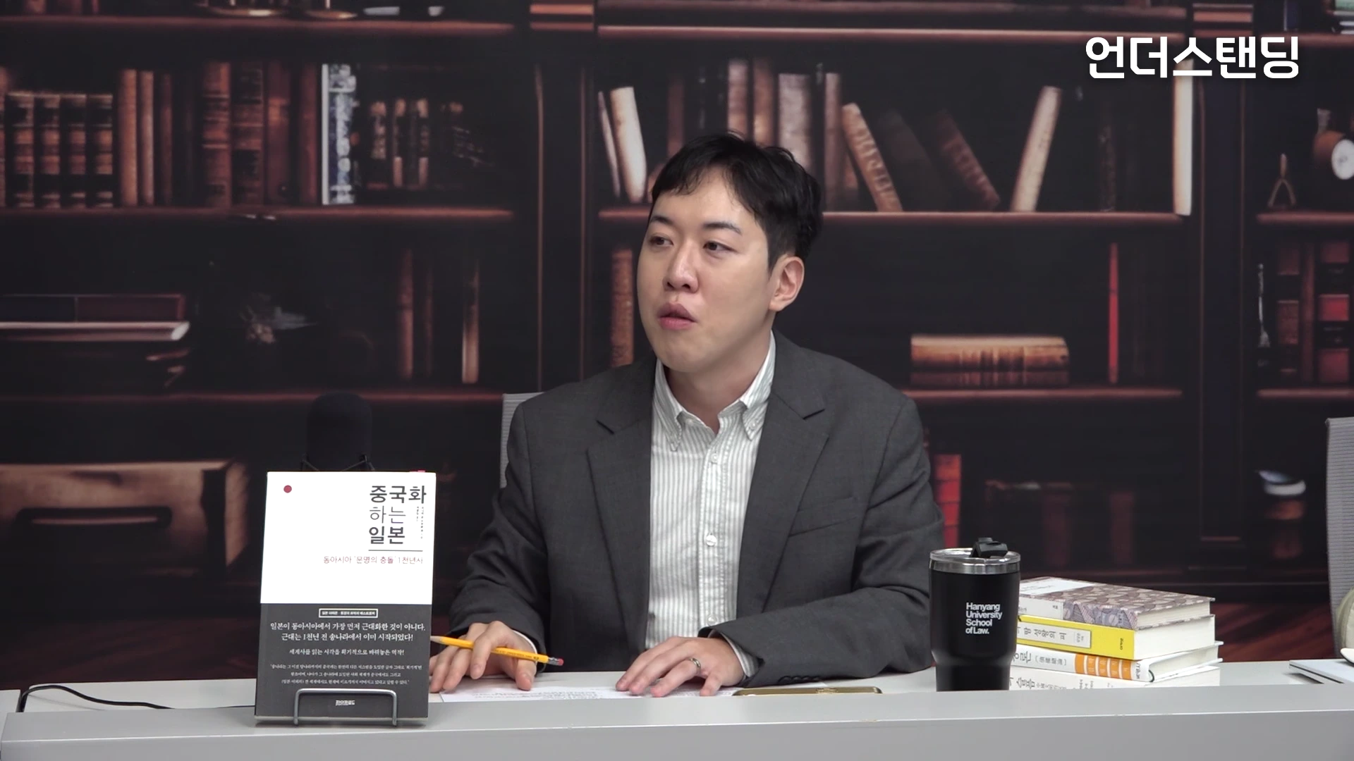 서재 배경의 스튜디오에서 재킷을 입은 남성이 테이블에 앉아 책을 앞에 두고 이야기를 나누고 있는 모습