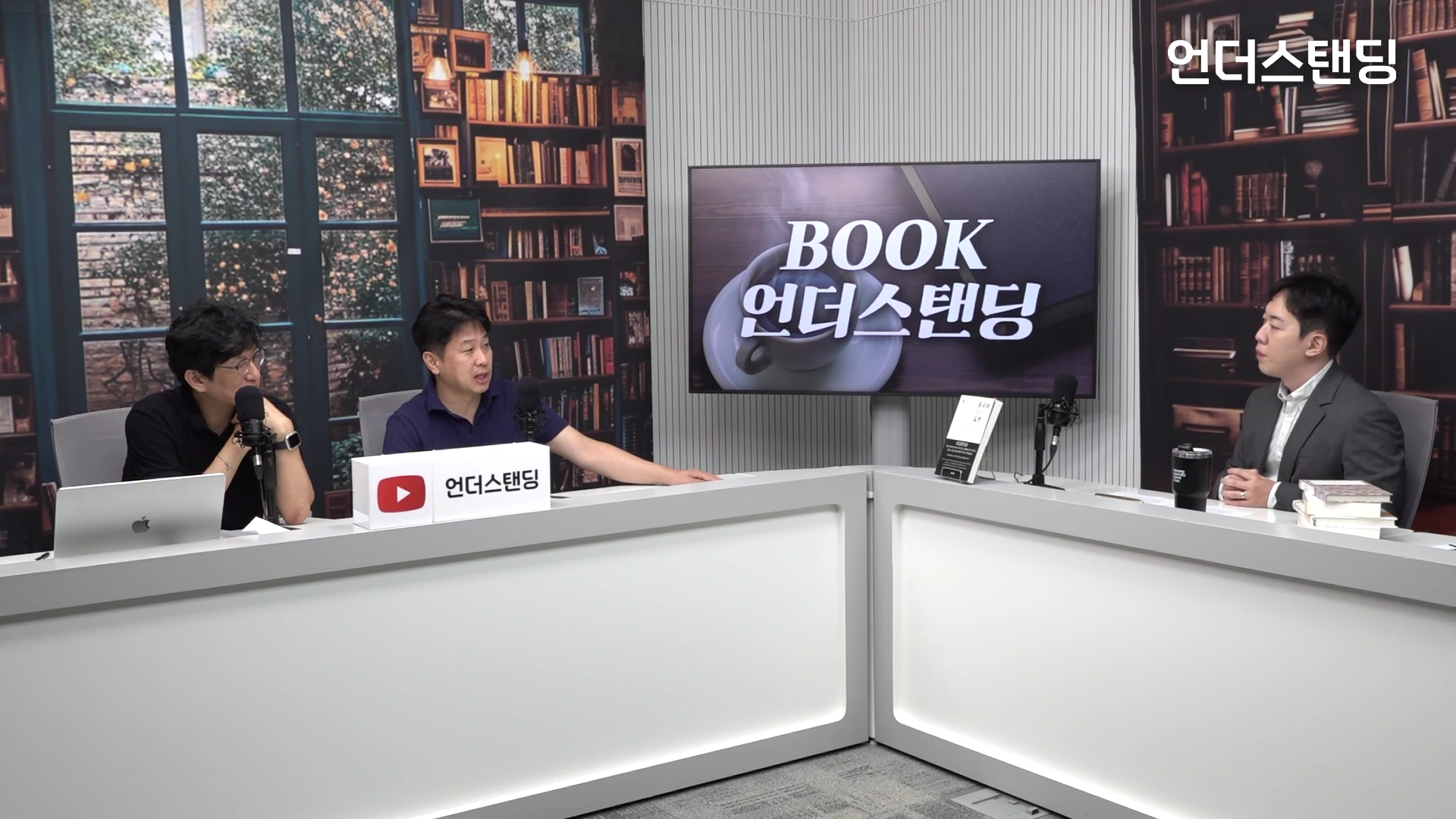 서재 배경의 스튜디오에서 세 명의 남성이 책상에 앉아 대화를 나누는 모습.