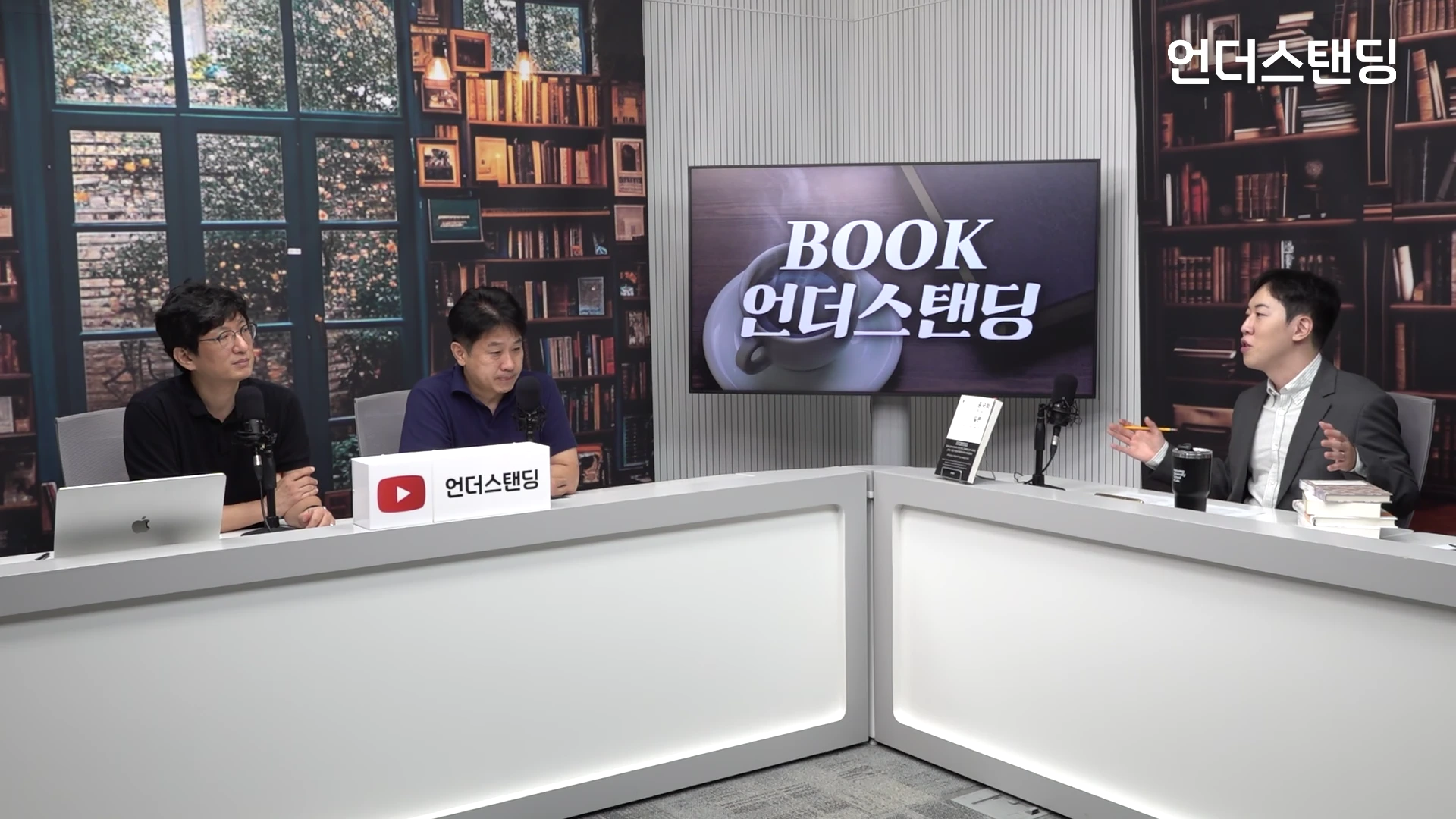 서재 배경의 스튜디오에서 세 명의 남성이 책상에 앉아 마이크를 앞에 두고 대화를 나누고 있는 모습입니다.