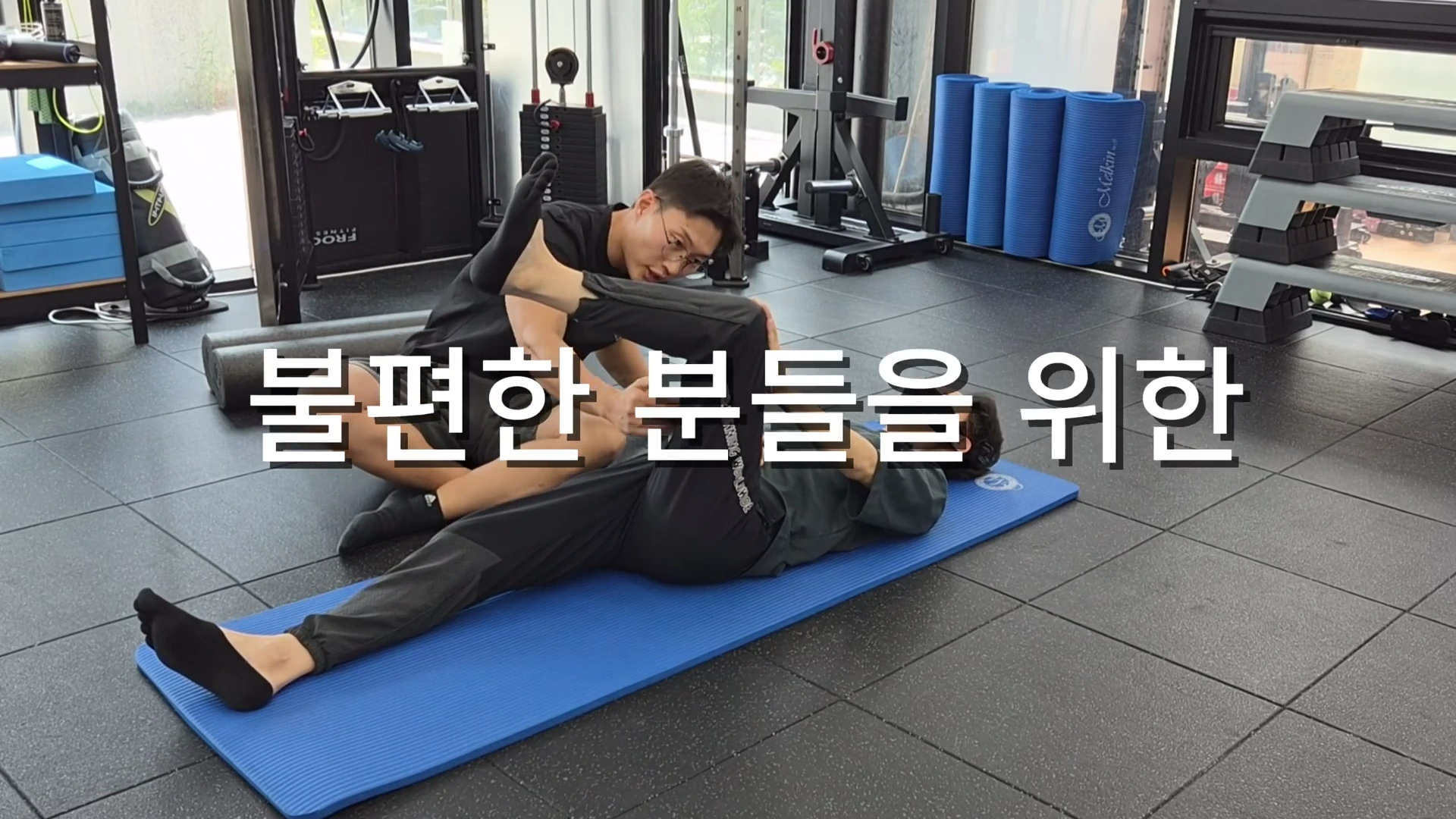 피트니스 센터 매트 위에서 한 남성이 누워 있는 다른 남성의 다리를 잡고 운동 동작을 가이드하고 있으며 화면 중앙에는 '불편한 분들을 위한'이라는 흰색 자막이 배치되어 있다.