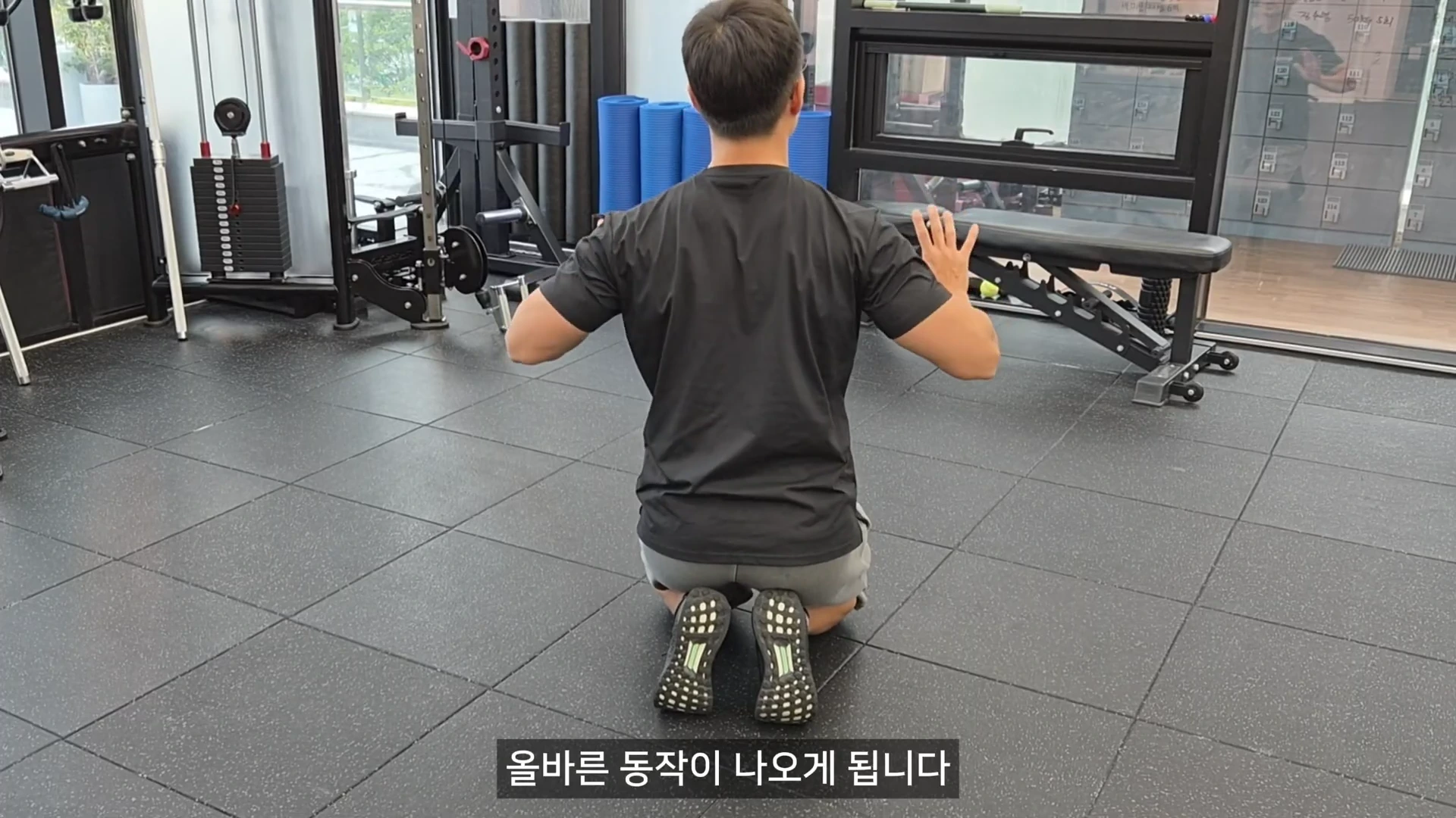 헬스장에서 검은색 티셔츠를 입은 남성이 무릎을 꿇고 앉아 푸시업 자세를 연습하며 팔을 벌리고 있는 뒷모습
