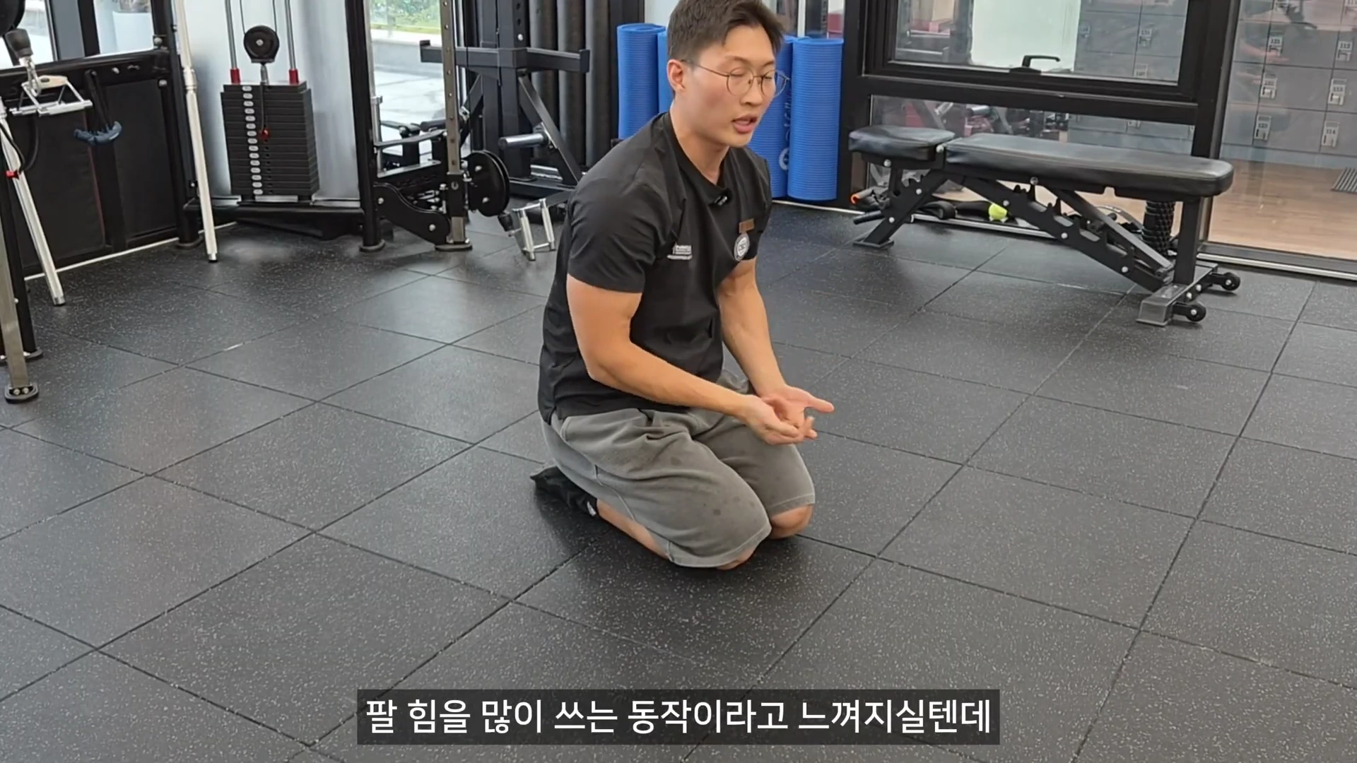 안경을 쓴 남성 트레이너가 체육관 바닥에 무릎을 꿇고 앉아 푸시업 동작에 대해 설명하고 있는 모습이다.